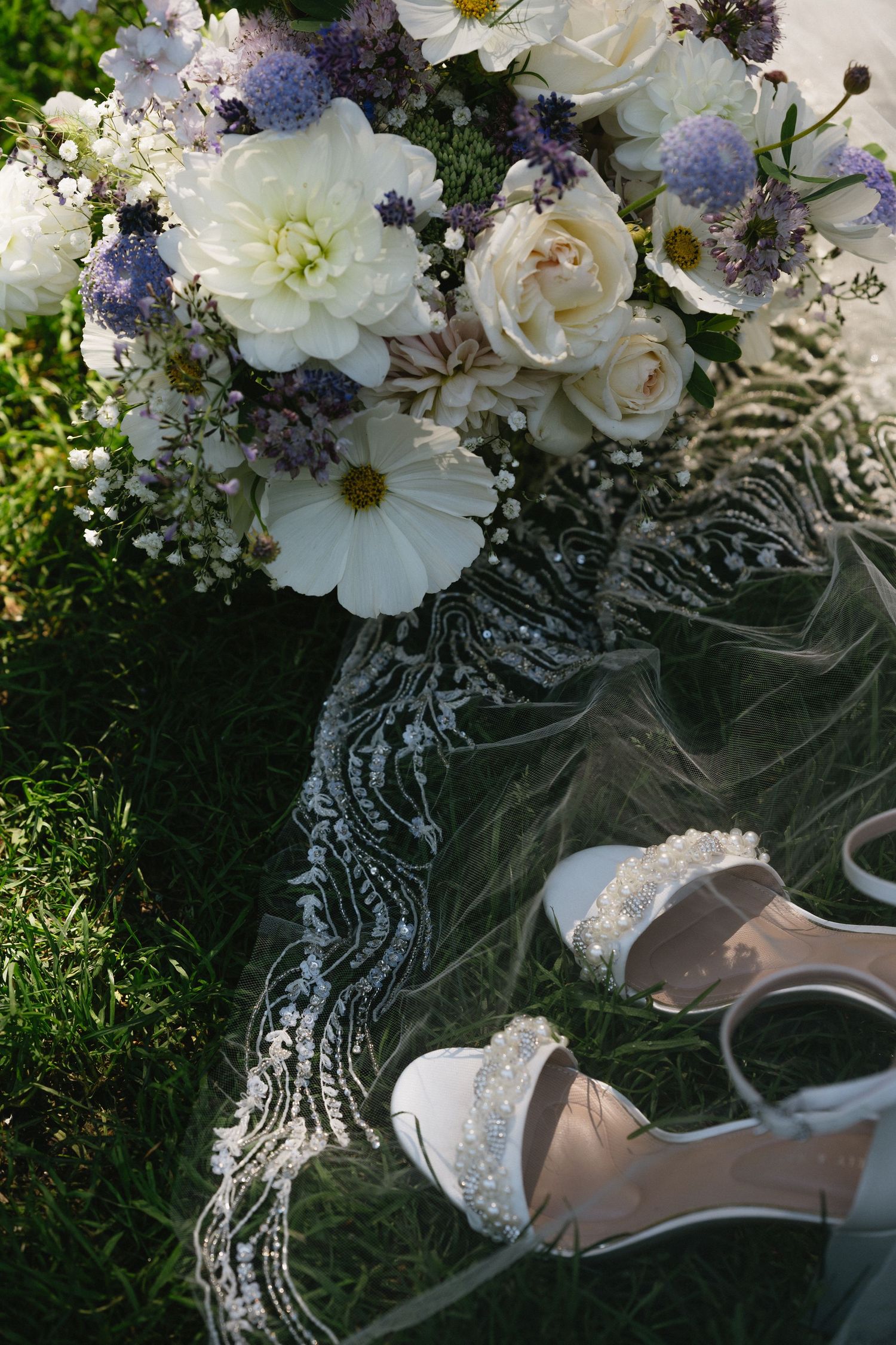 White and purple bridal bouquet with lace veil and pearl-adorned heels on green grass.