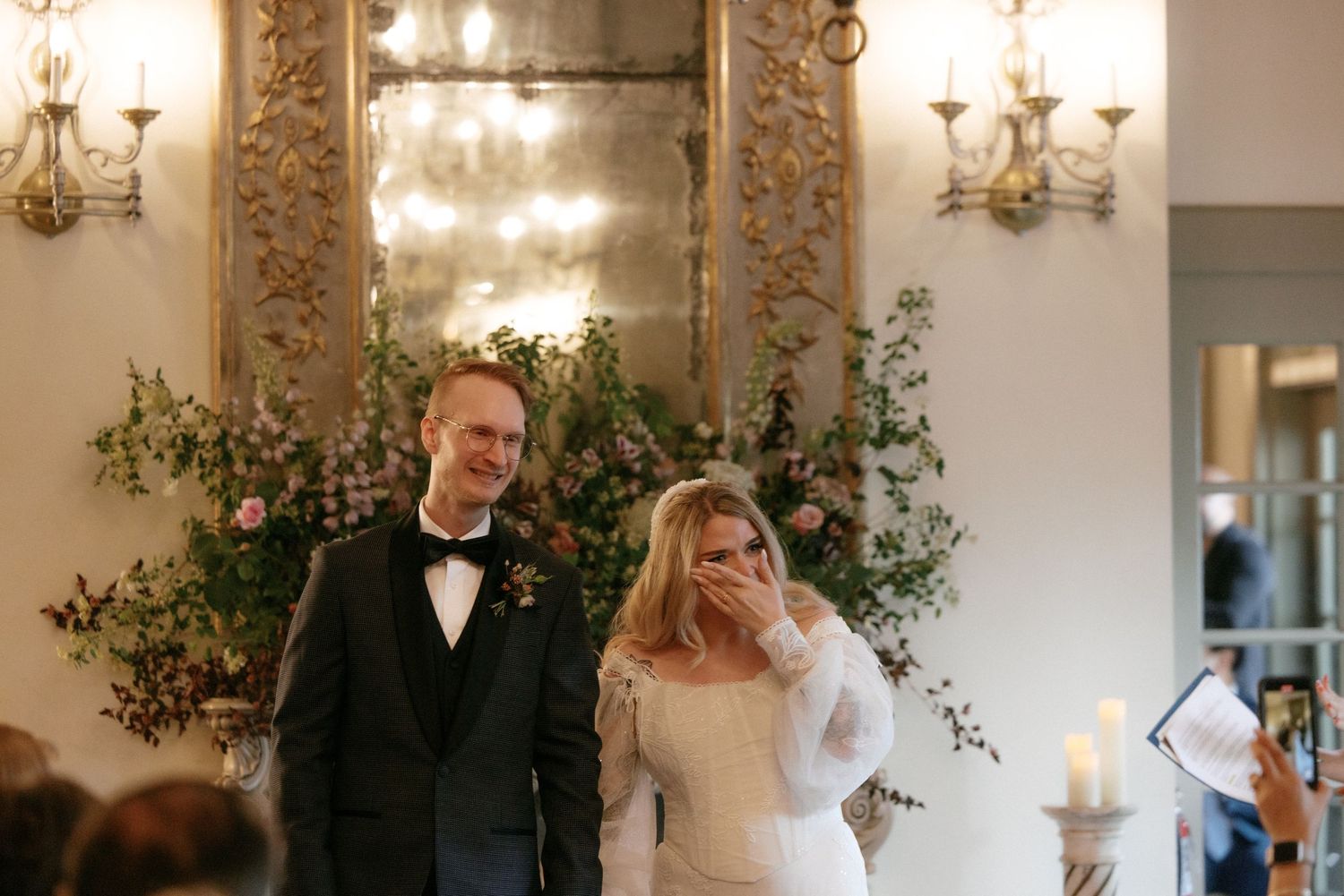 Newlyweds smile joyfully at guests before ornate mirror and greenery at elegant wedding ceremony.