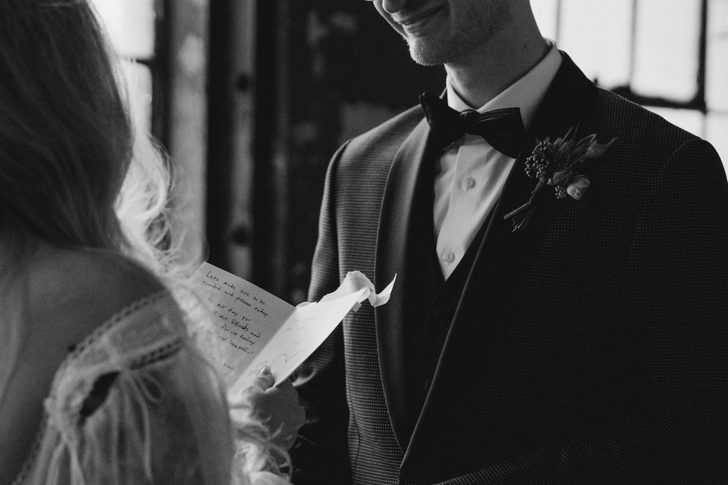 Groom smiles as bride reads heartfelt wedding vows in black and white photo.