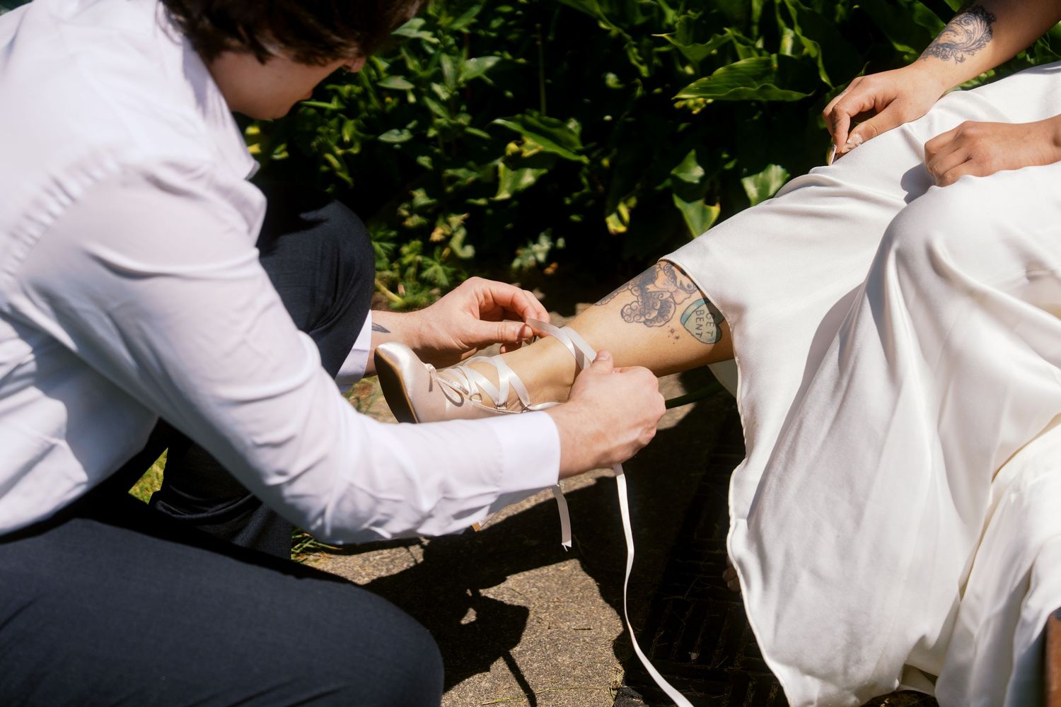 Wedding assistant helping bride with decorative garter on tattooed leg outdoors.
