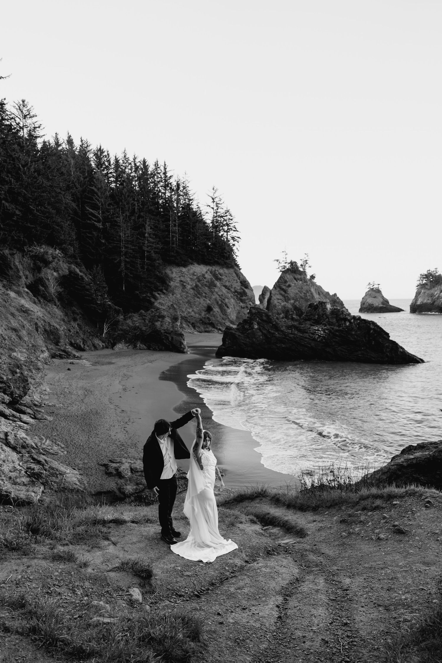 Black and white photo of groom twirling bride on a scenic coastal beach with sea stacks.
