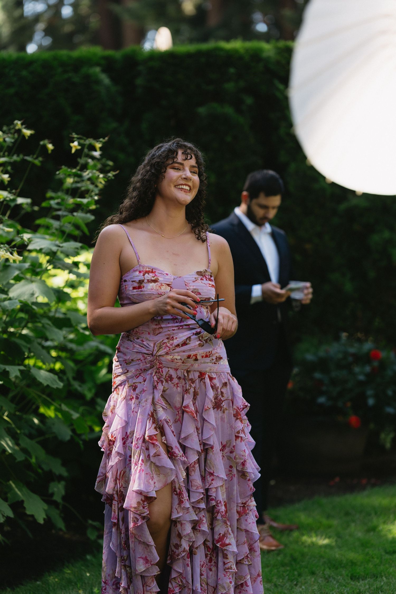 Woman in floral ruffled pink dress smiling outdoors at wedding reception in garden setting.