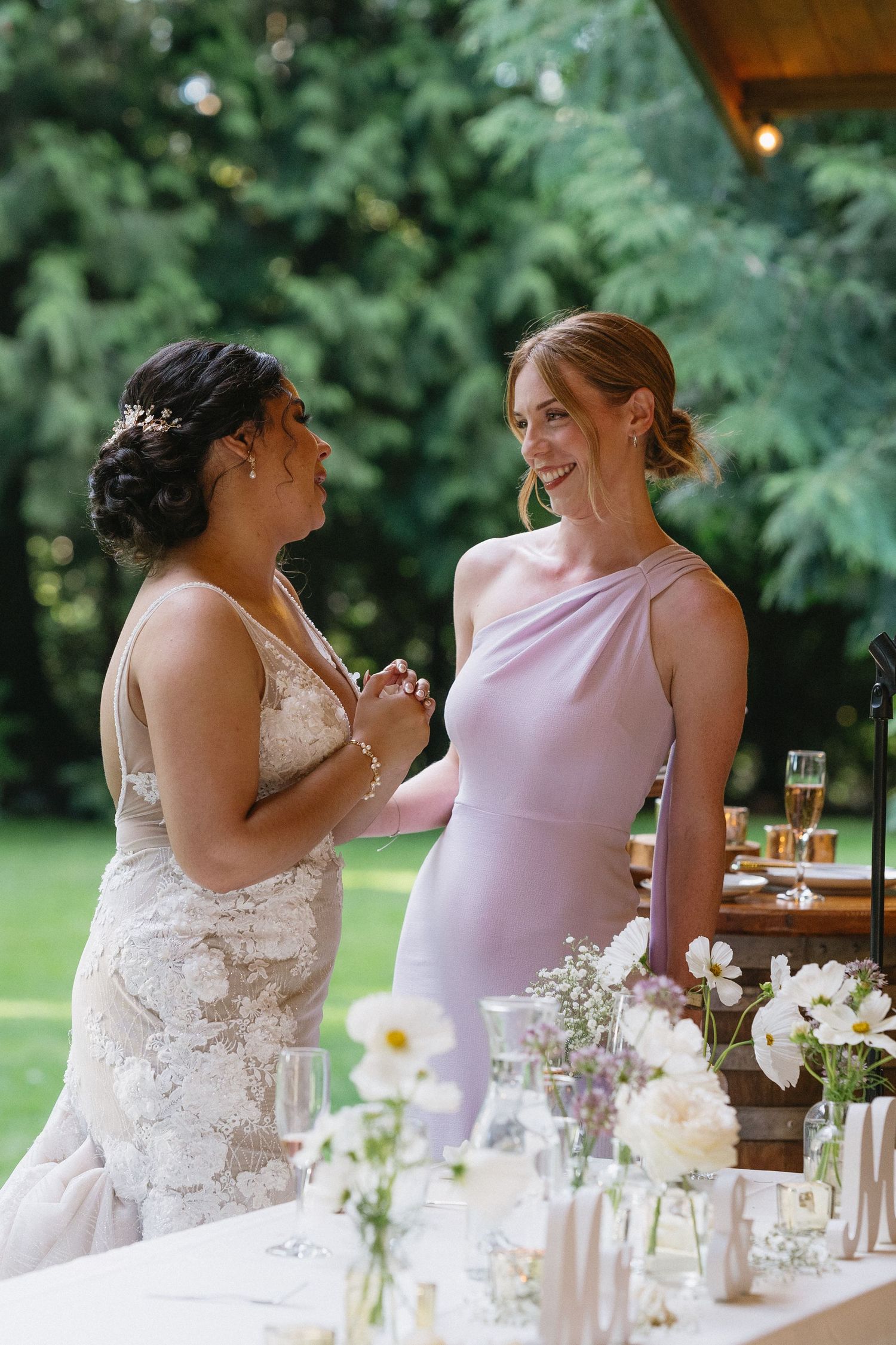 Bride in lace gown sharing a joyful moment with bridesmaid in lavender dress at wedding reception.