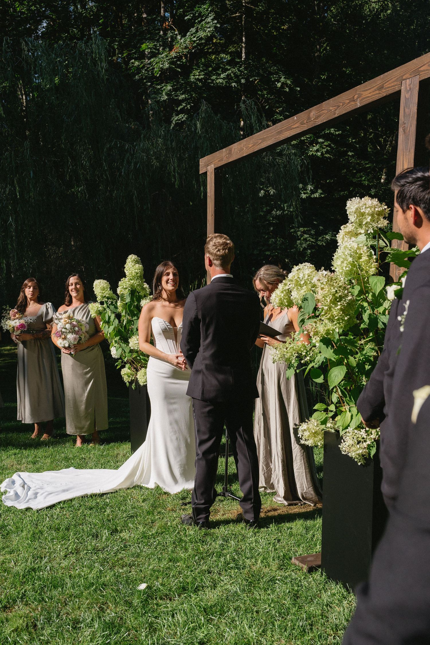 Bride and groom exchange vows at outdoor ceremony beneath wooden arch adorned with hydrangeas and greenery.