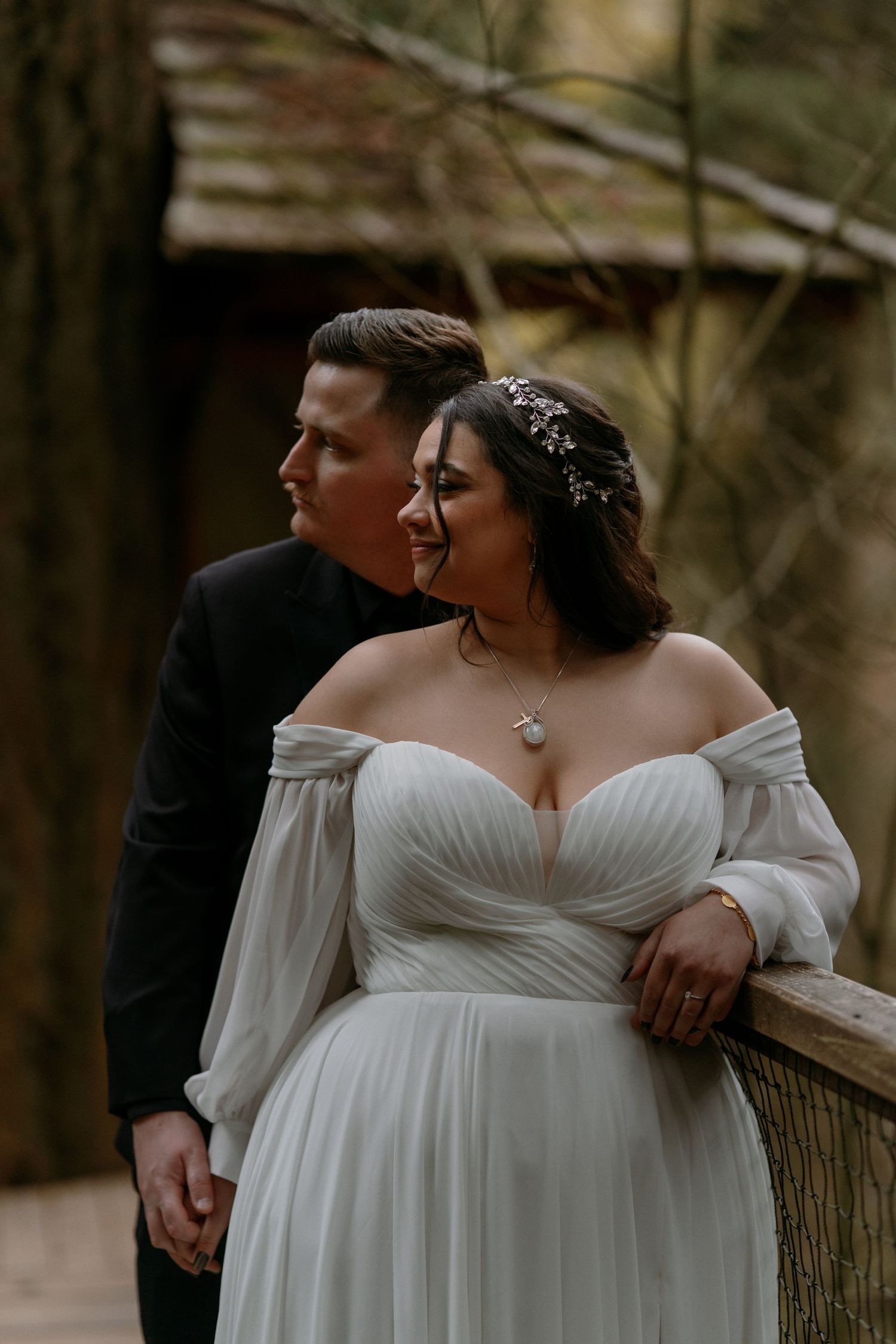Elegant bride in off-shoulder white gown with groom in black suit posing at an outdoor forest wedding.