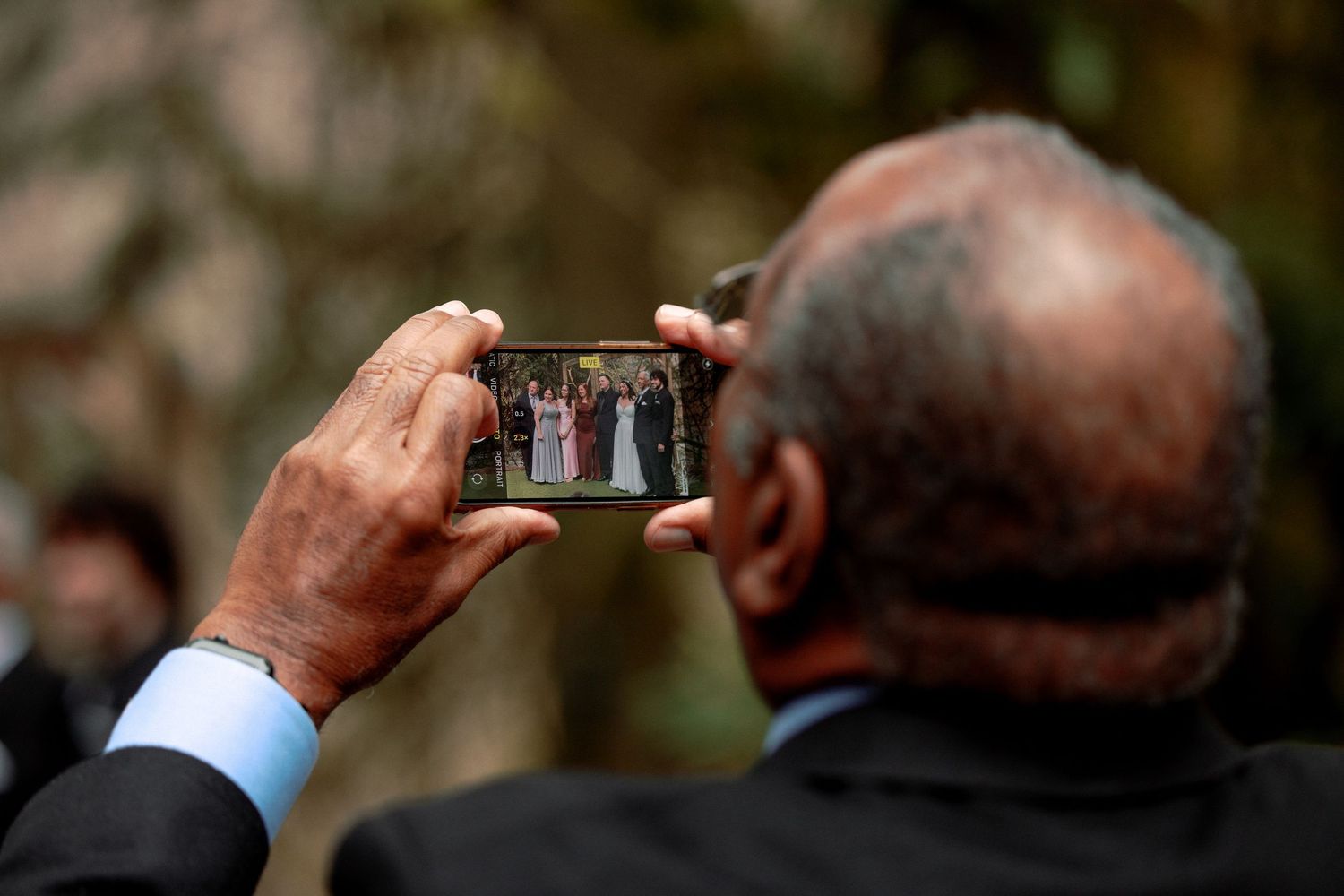 Wedding guest photographing bridal party on smartphone at an outdoor forest wedding ceremony.