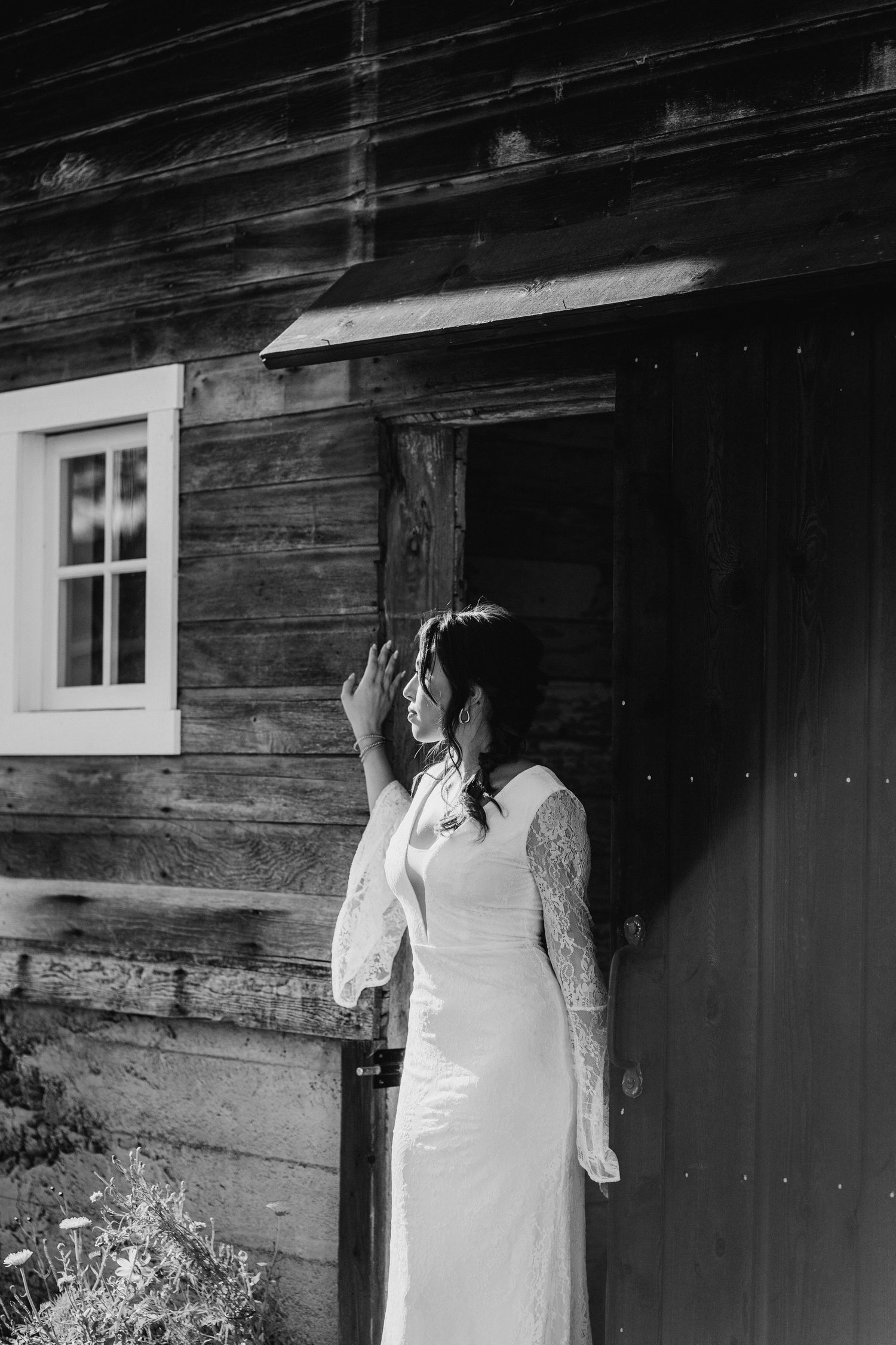 Black and white photo of bride in lace wedding dress leaning against rustic wooden barn.