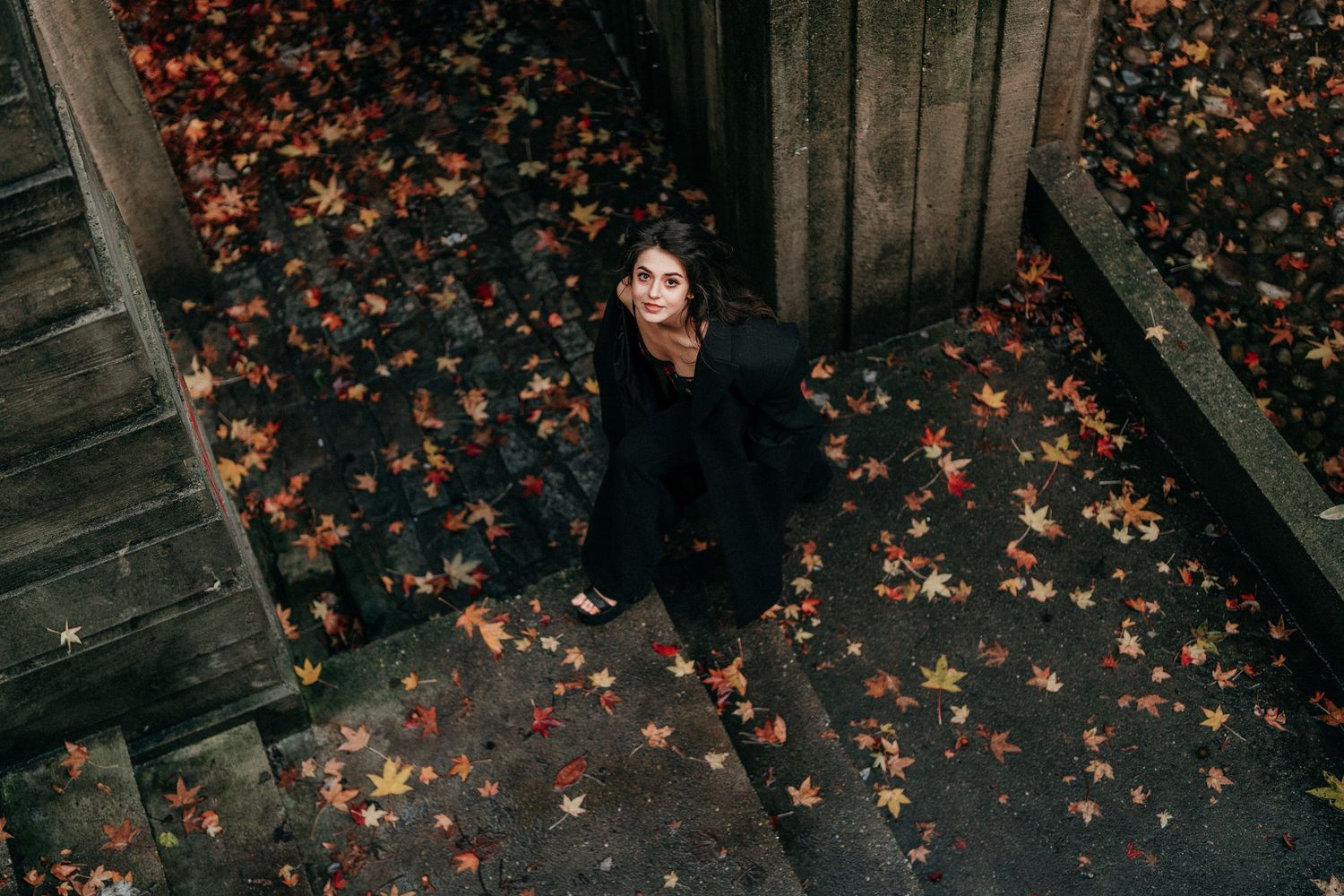 Overhead view of woman in black dress sitting on steps surrounded by colorful fallen autumn leaves.