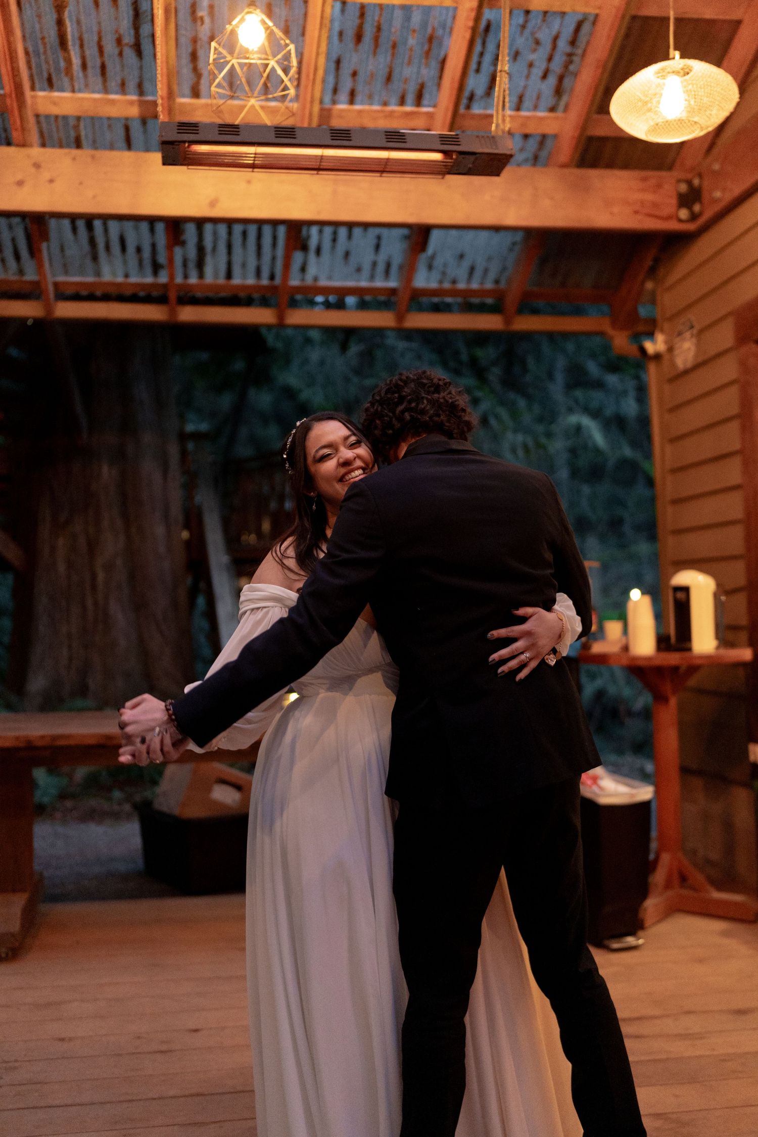 Smiling bride in an off-shoulder white gown embraces groom while dancing on a wooden deck at their outdoor wedding.
