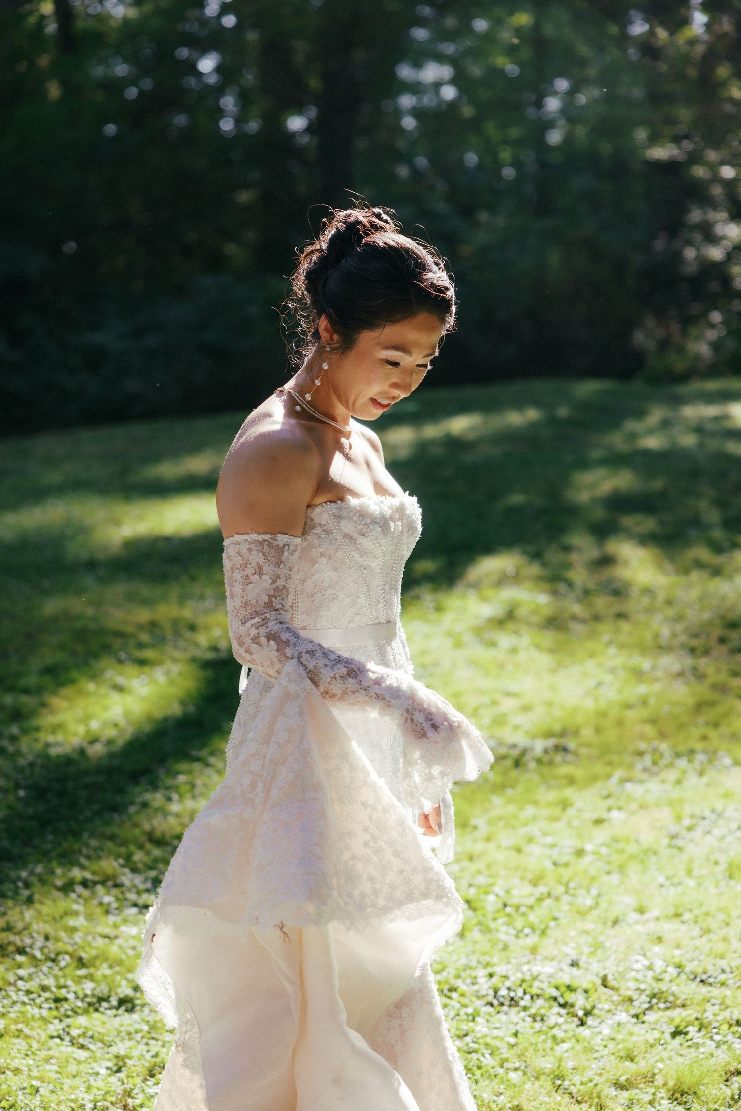 Elegant bride in strapless lace wedding gown with sheer gloves twirls on sunlit green grass in a forest.
