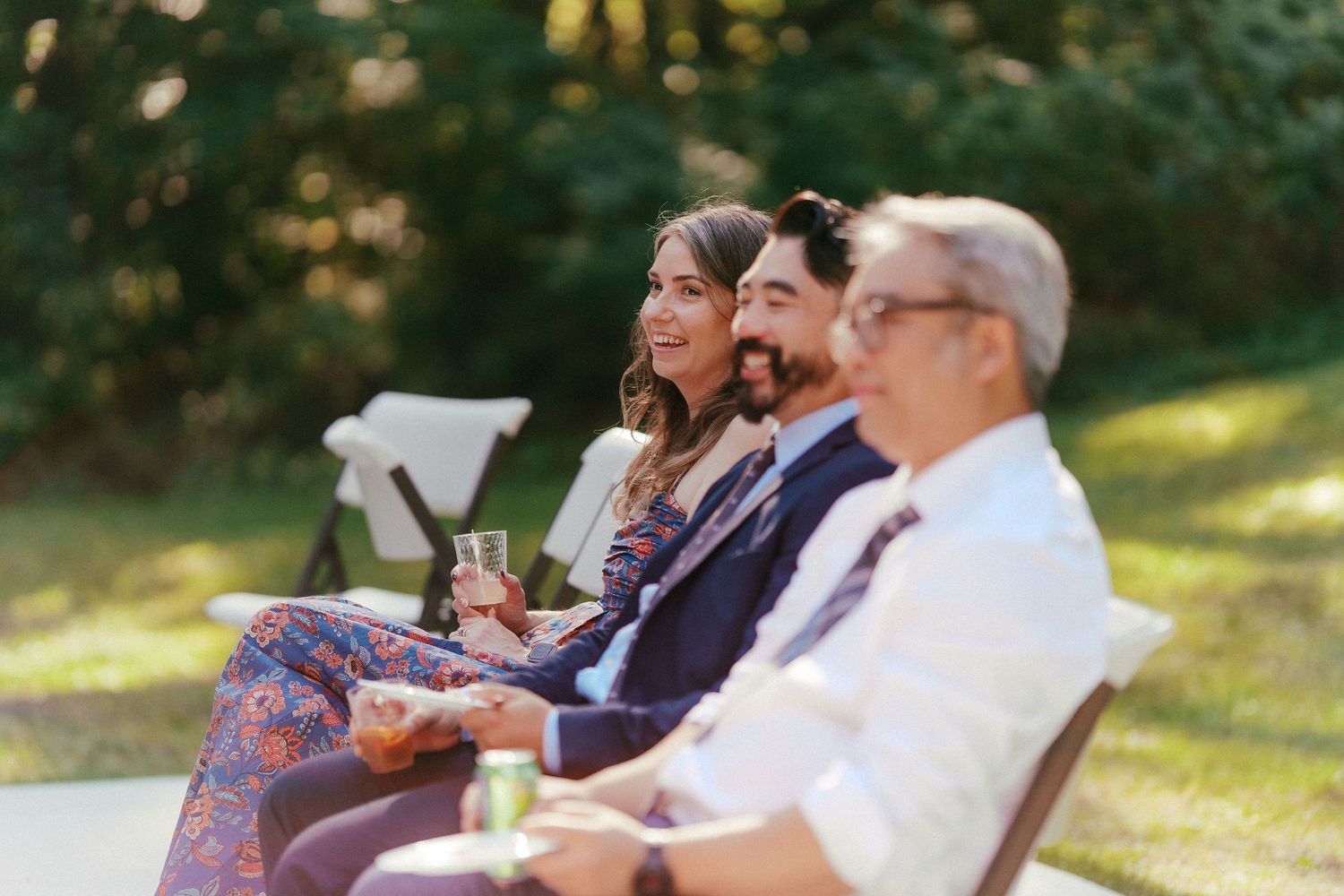 Wedding guests smile and relax in chairs outdoors, holding drinks during a sunlit garden ceremony.