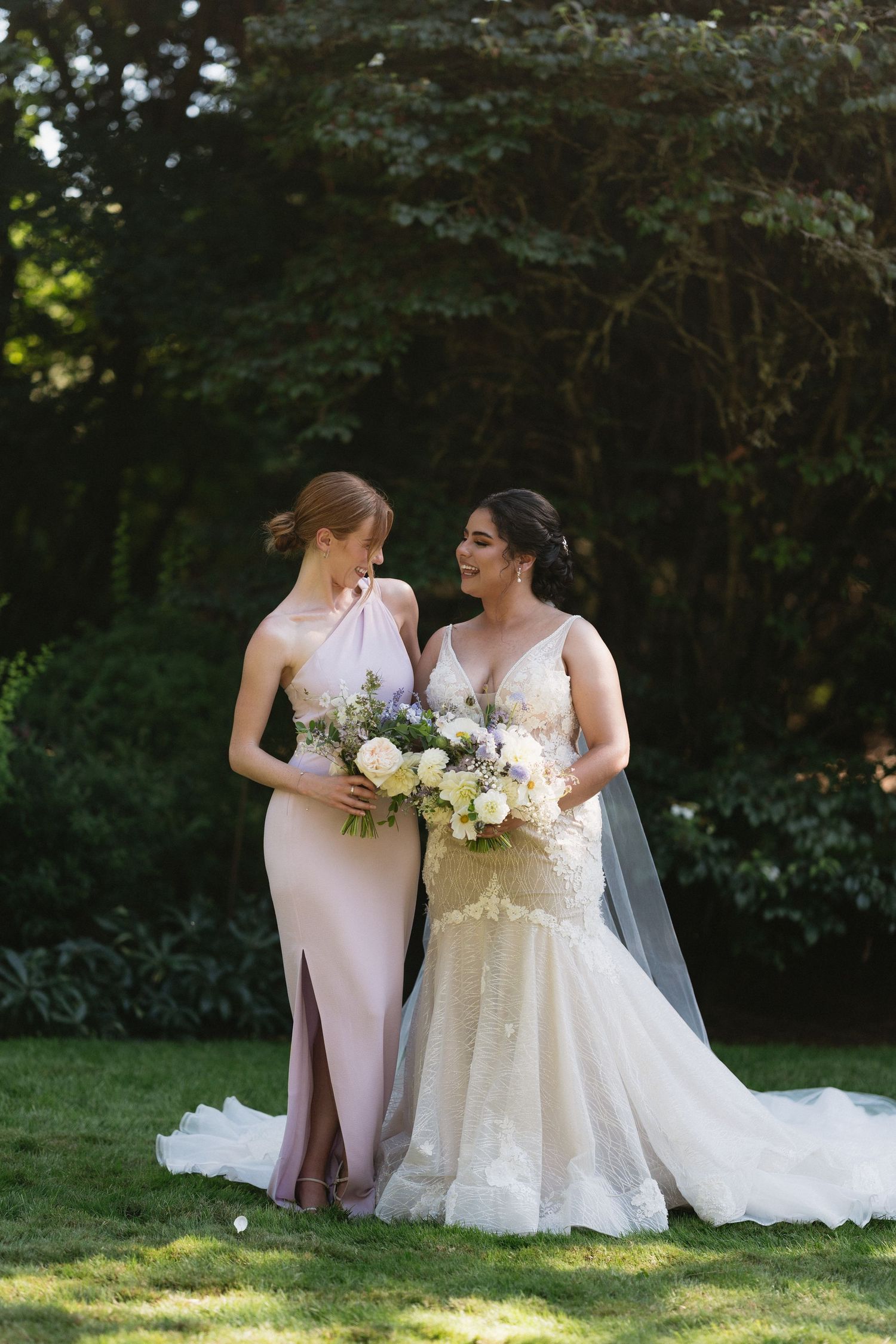 Bride in lace gown and bridesmaid in blush pink dress holding bouquets in a lush garden setting.