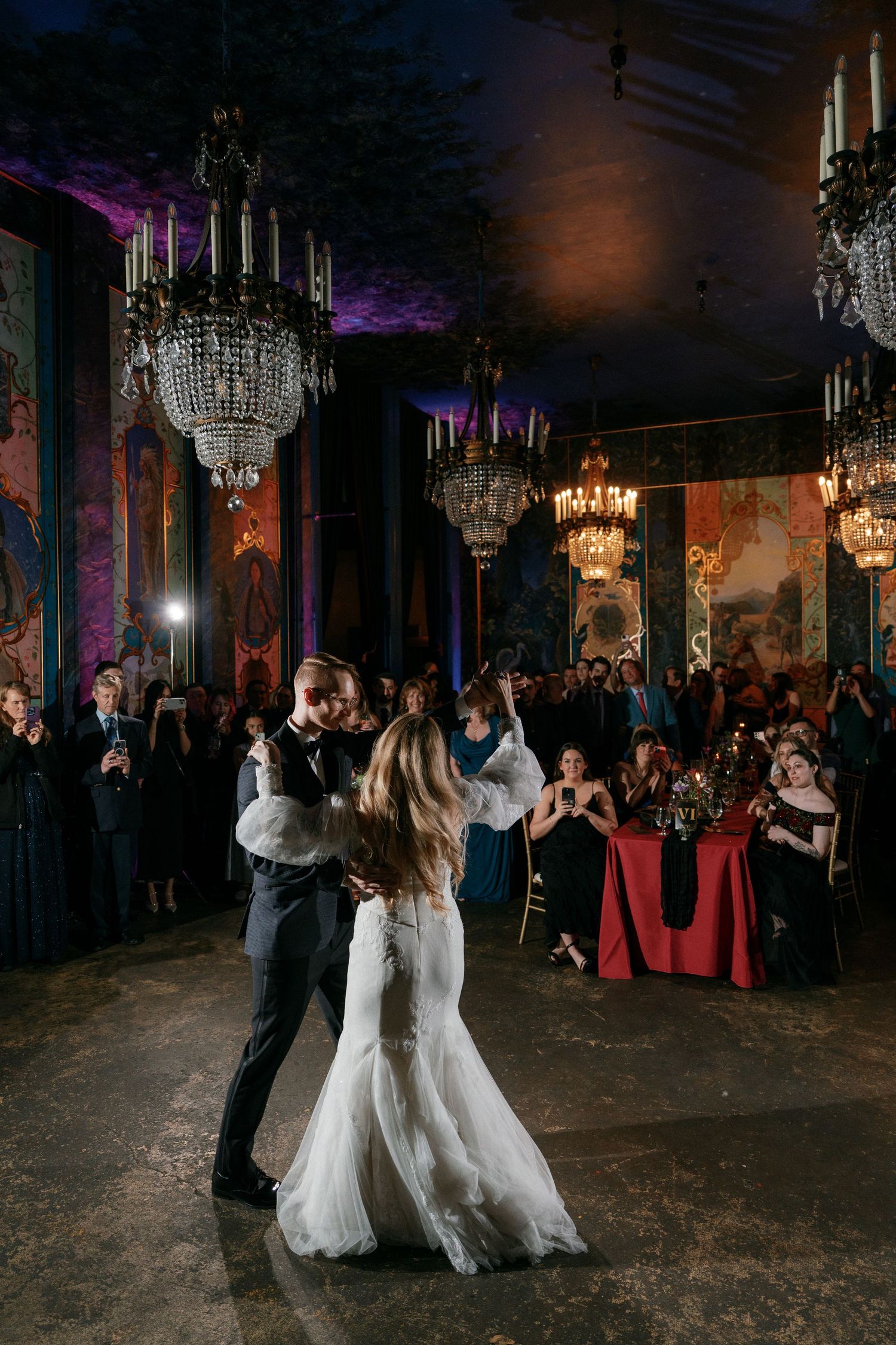 Couple dancing at wedding reception in ornate ballroom with crystal chandeliers.