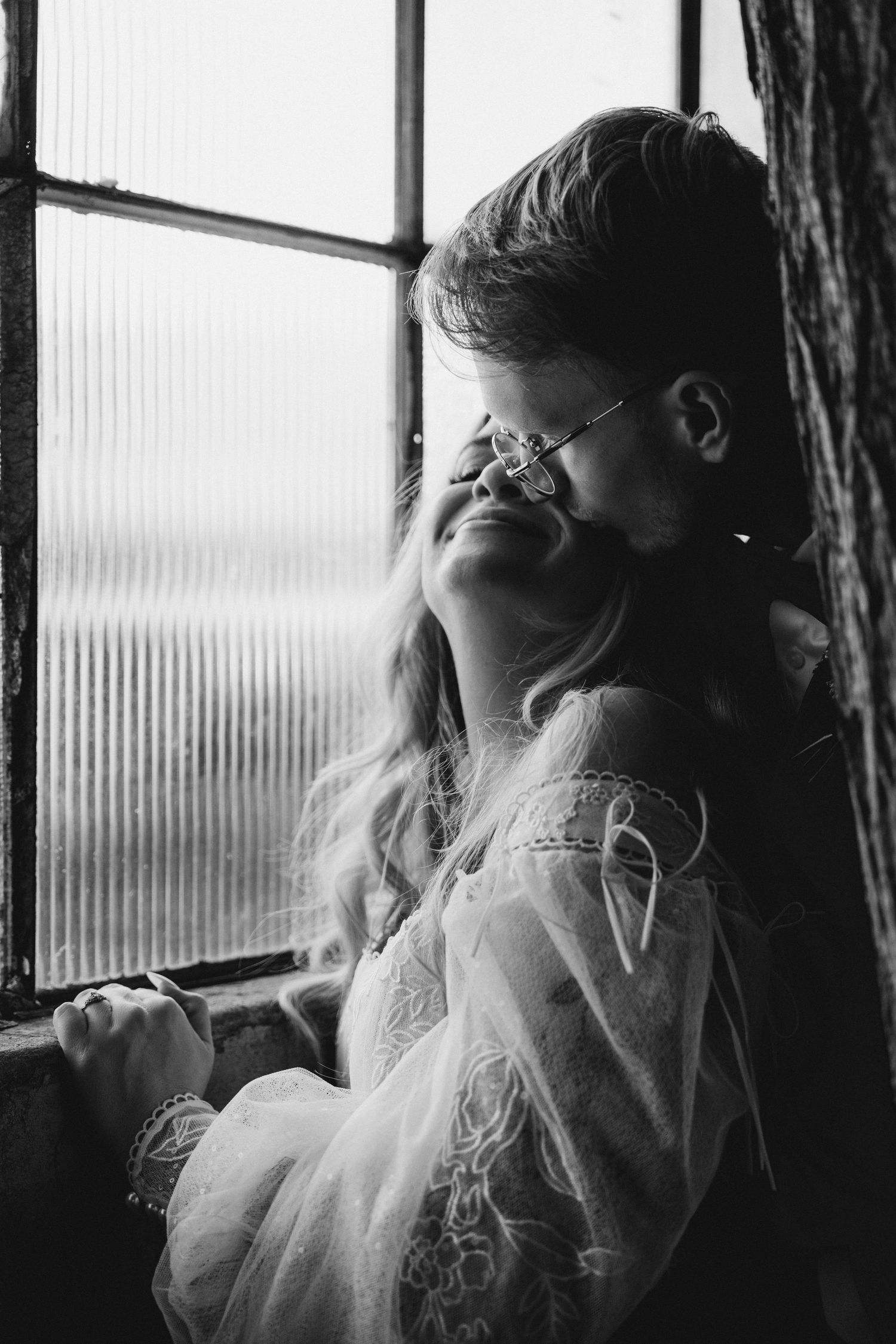Black and white portrait of smiling bride being kissed by groom wearing glasses near a vintage window with soft light.