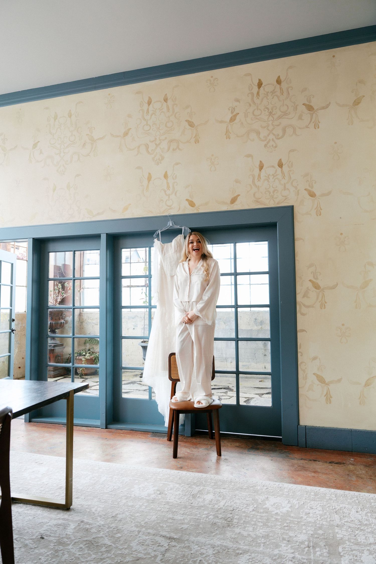 Bride in white robe standing on chair holding wedding dress in elegant decorated room.
