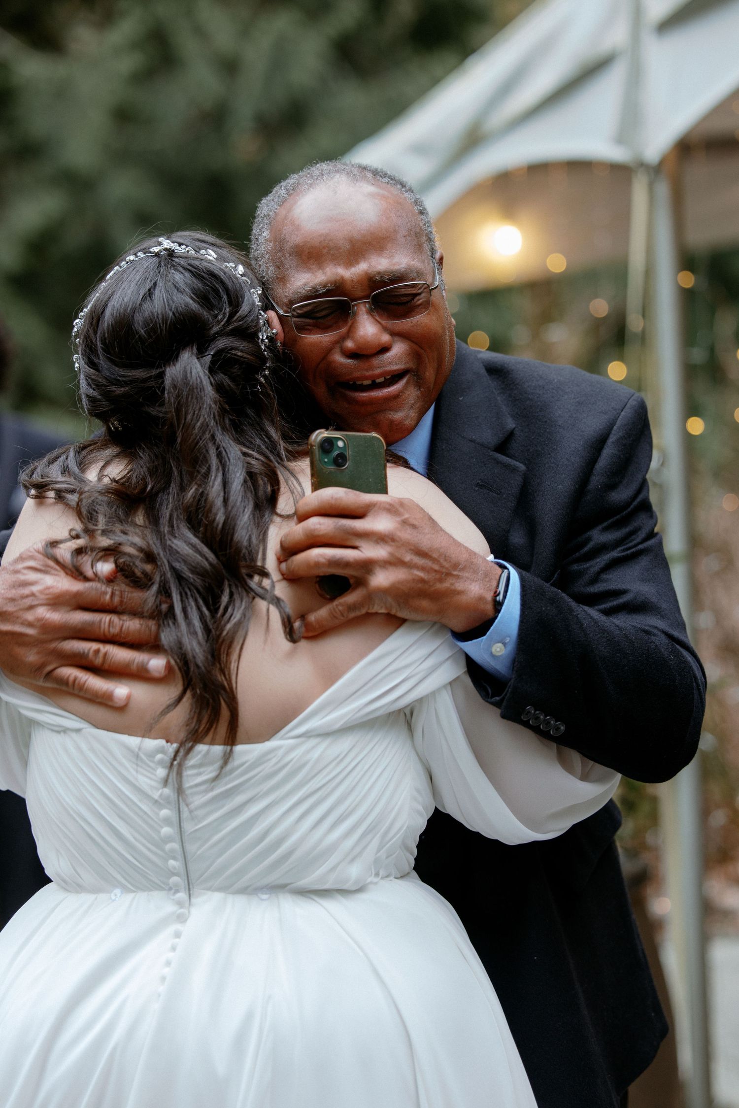 Emotional older man hugging bride in white wedding gown at outdoor reception celebration.