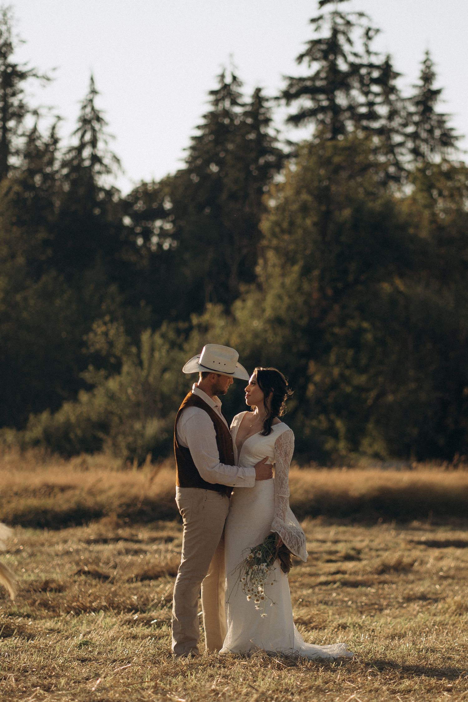 Bride and groom share a romantic moment in a golden autumn field surrounded by tall evergreen trees.
