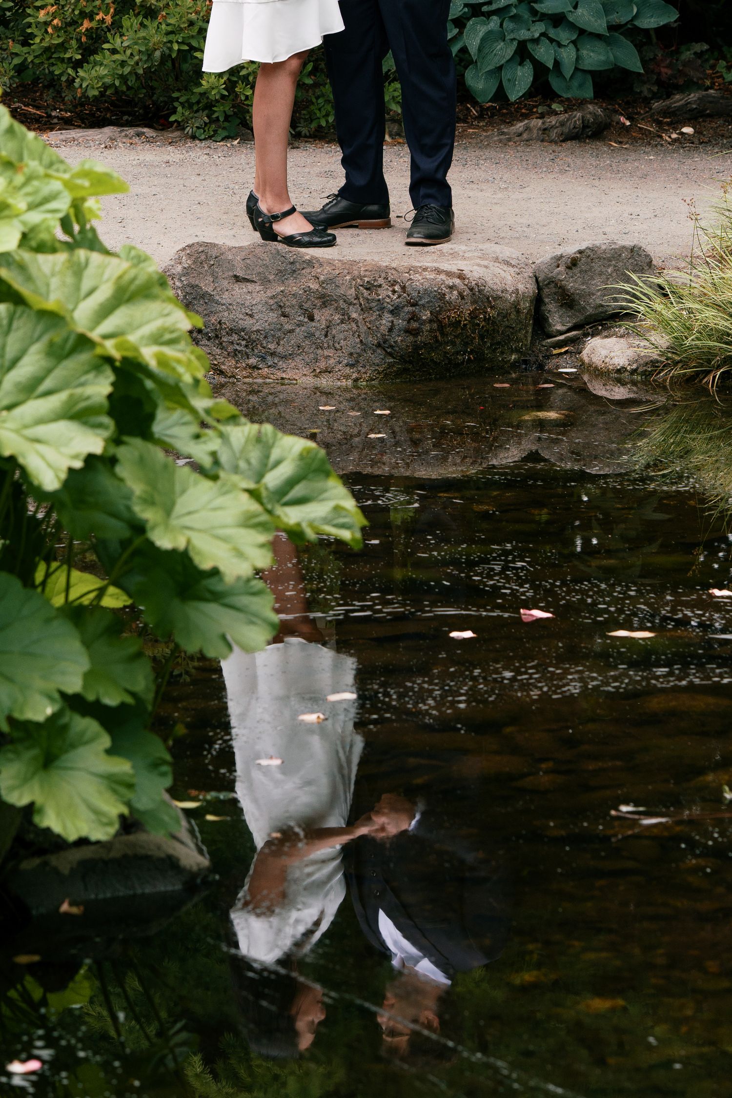 Couple's feet reflected in a garden pond, woman in black shoes and man in dress shoes standing on a rock.