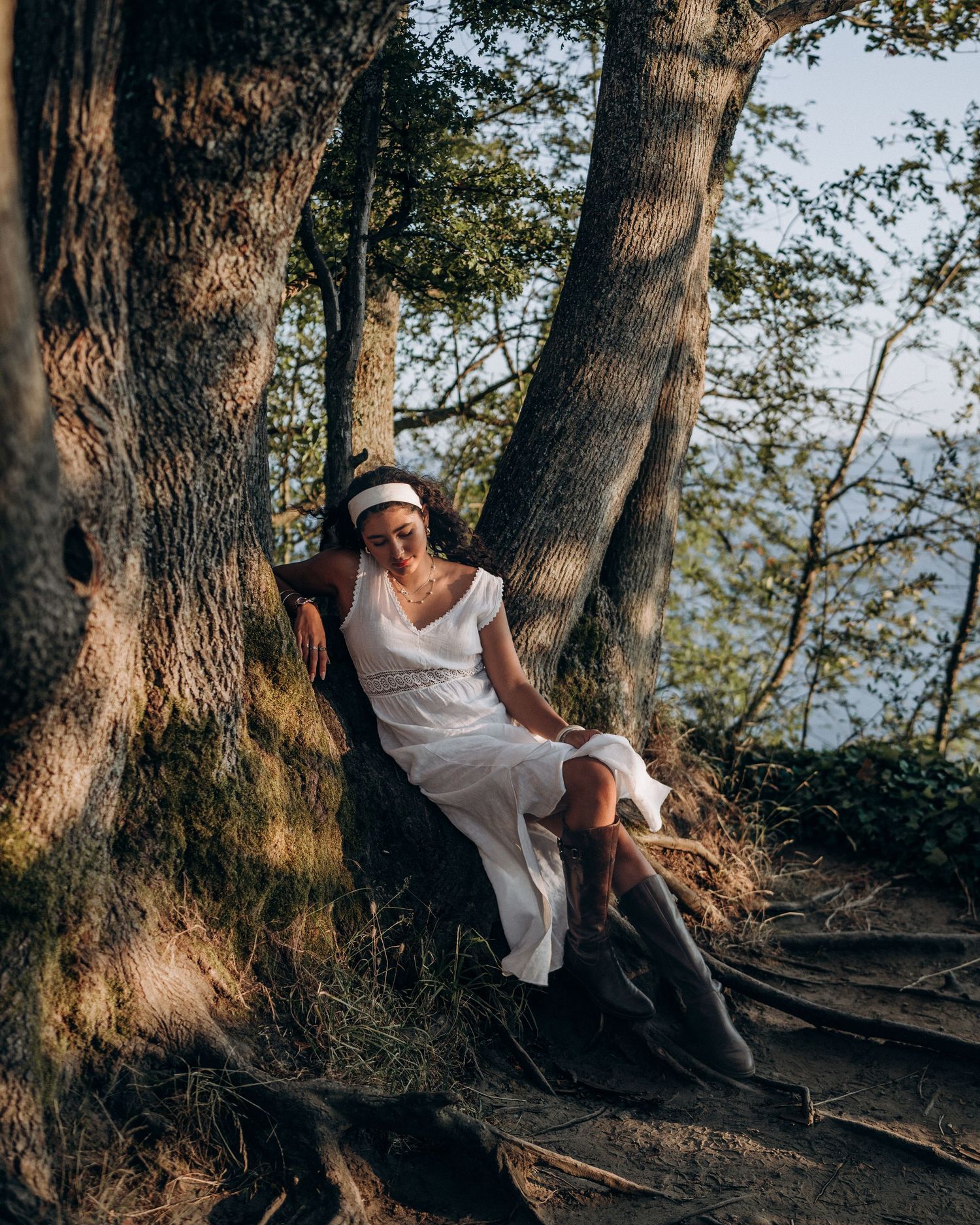 Woman in white dress and hat sitting against tree roots with scenic water view in background.