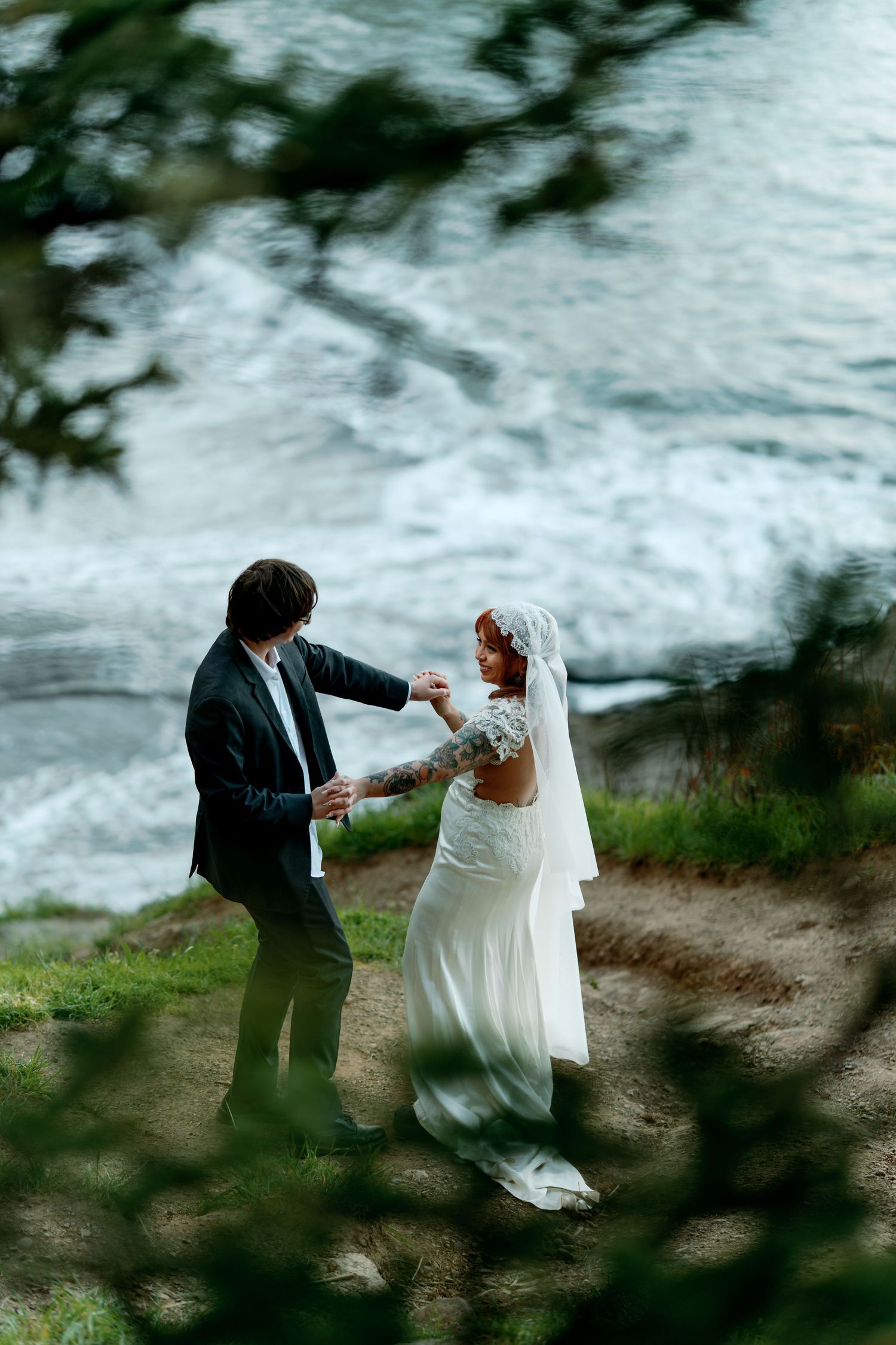 Bride and groom dancing near ocean cliffs, bride in lace wedding dress with veil, groom in dark suit.