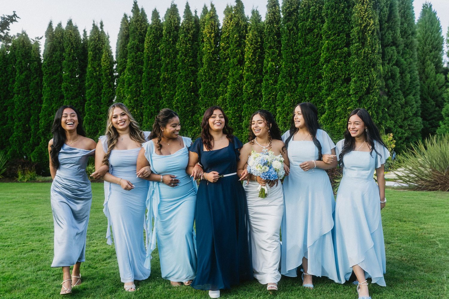 Six bridesmaids in light blue dresses laugh and walk together on green lawn.