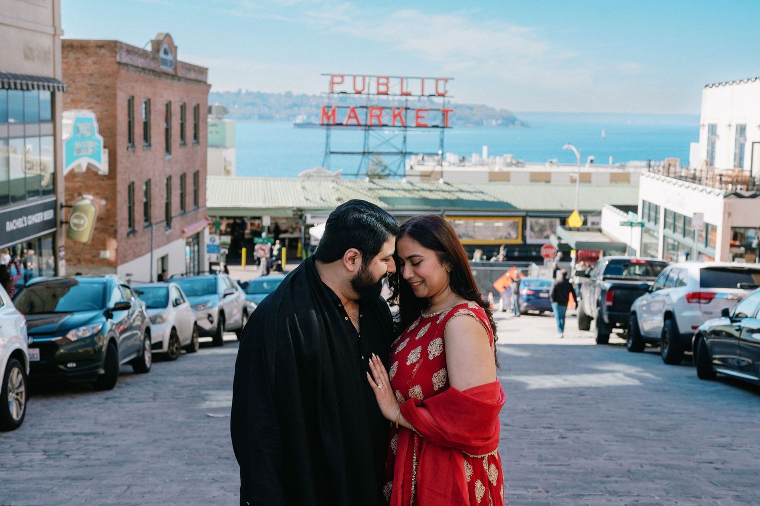 Couple in traditional Indian outfits sharing a romantic moment near Seattle's iconic Pike Place Market sign.