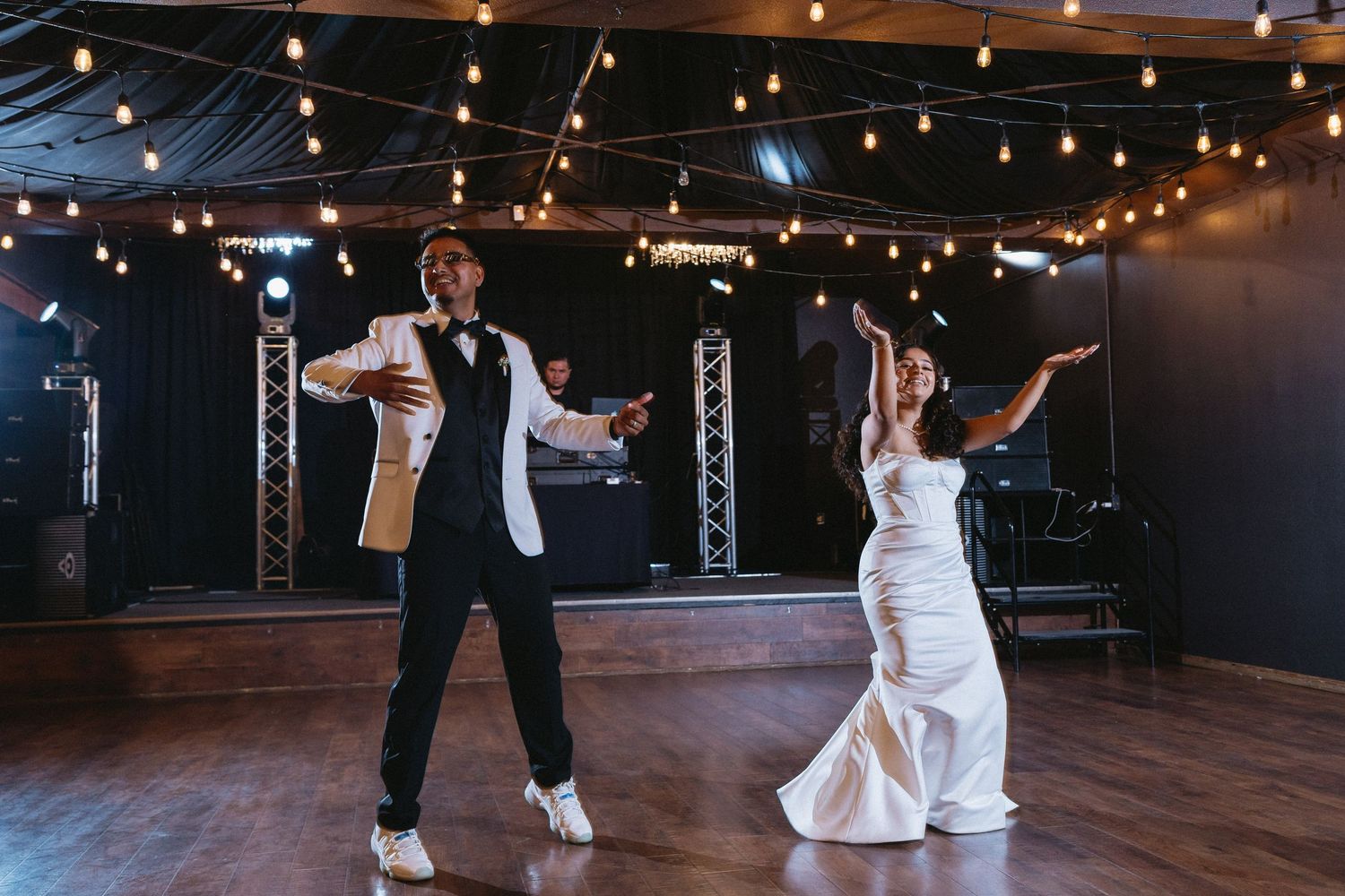 Bride and groom perform a fun choreographed wedding dance on a hardwood floor under string lights.