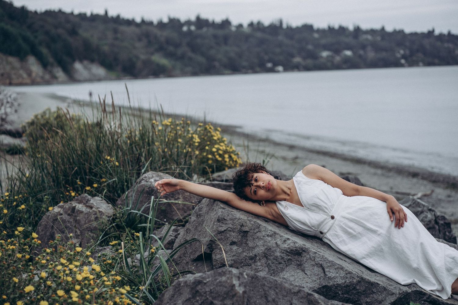Woman in white dress lies stretched across coastal rocks with yellow wildflowers nearby.