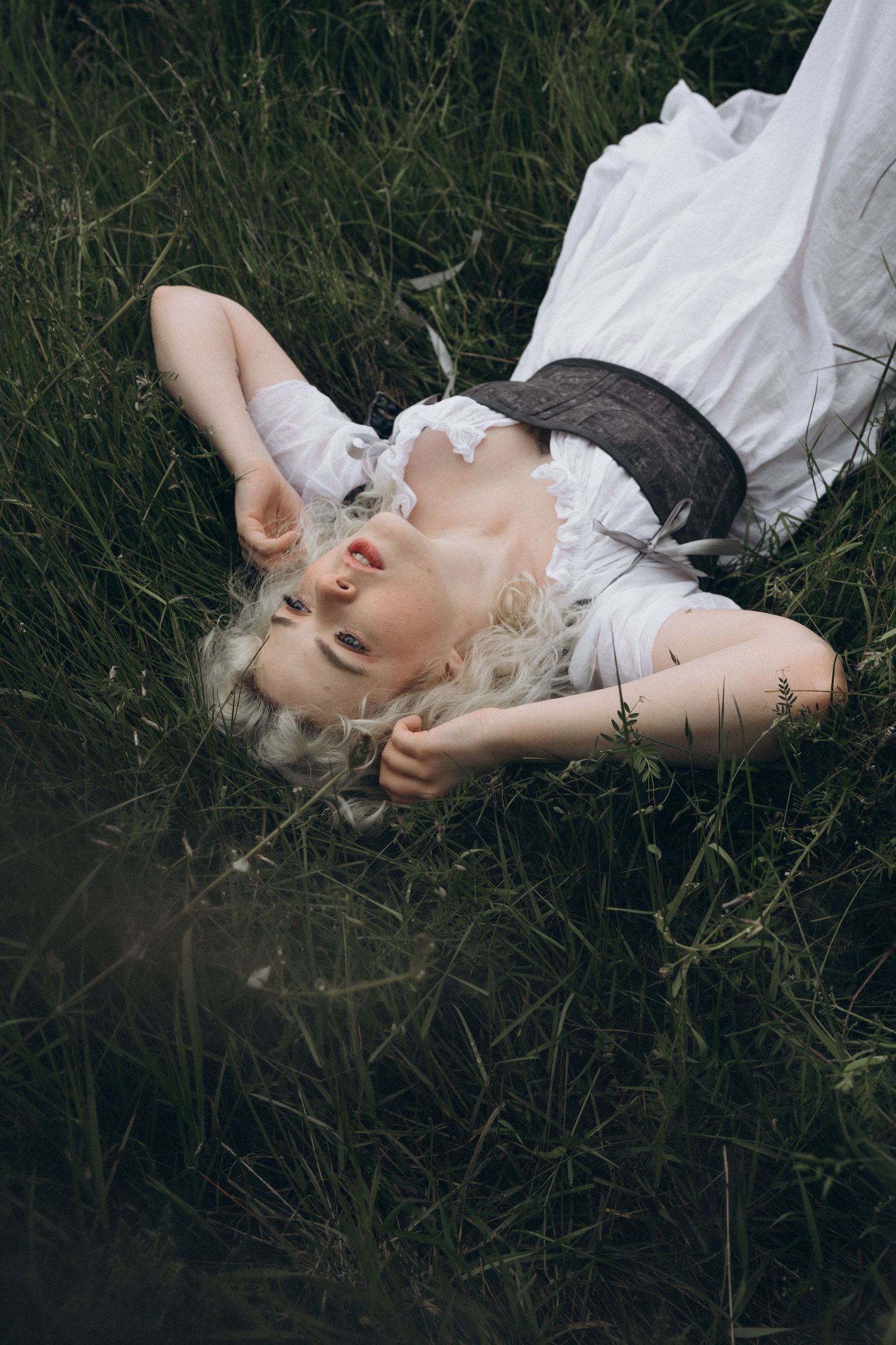 A blonde woman in a white dress and dark corset lies peacefully in tall green grass, gazing upward.