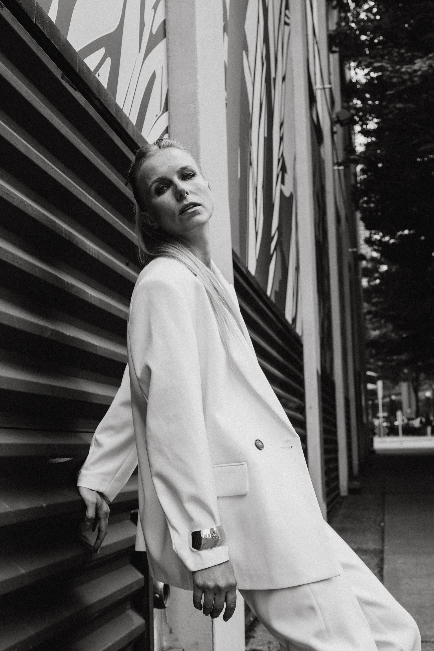 Woman in white oversized blazer and pants leans against corrugated metal wall in black and white urban fashion photo.