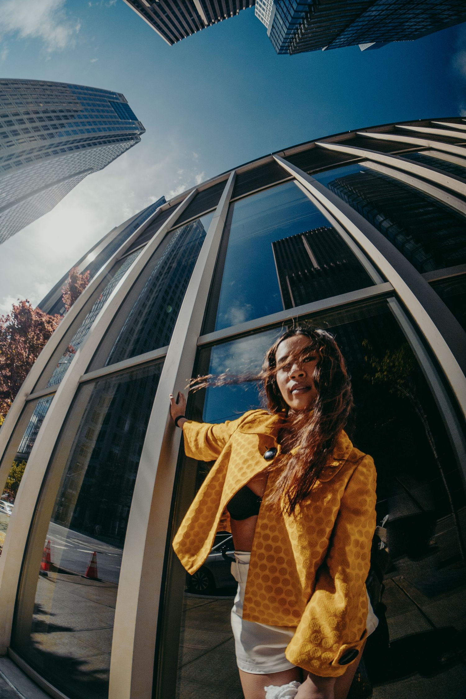 Woman in yellow patterned jacket leans against a curved glass building with tall skyscrapers under blue sky.