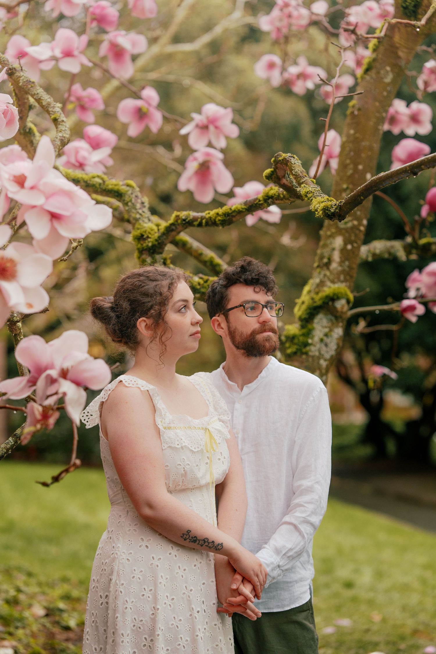 Couple holding hands under pink magnolia blossoms in a lush garden during spring.