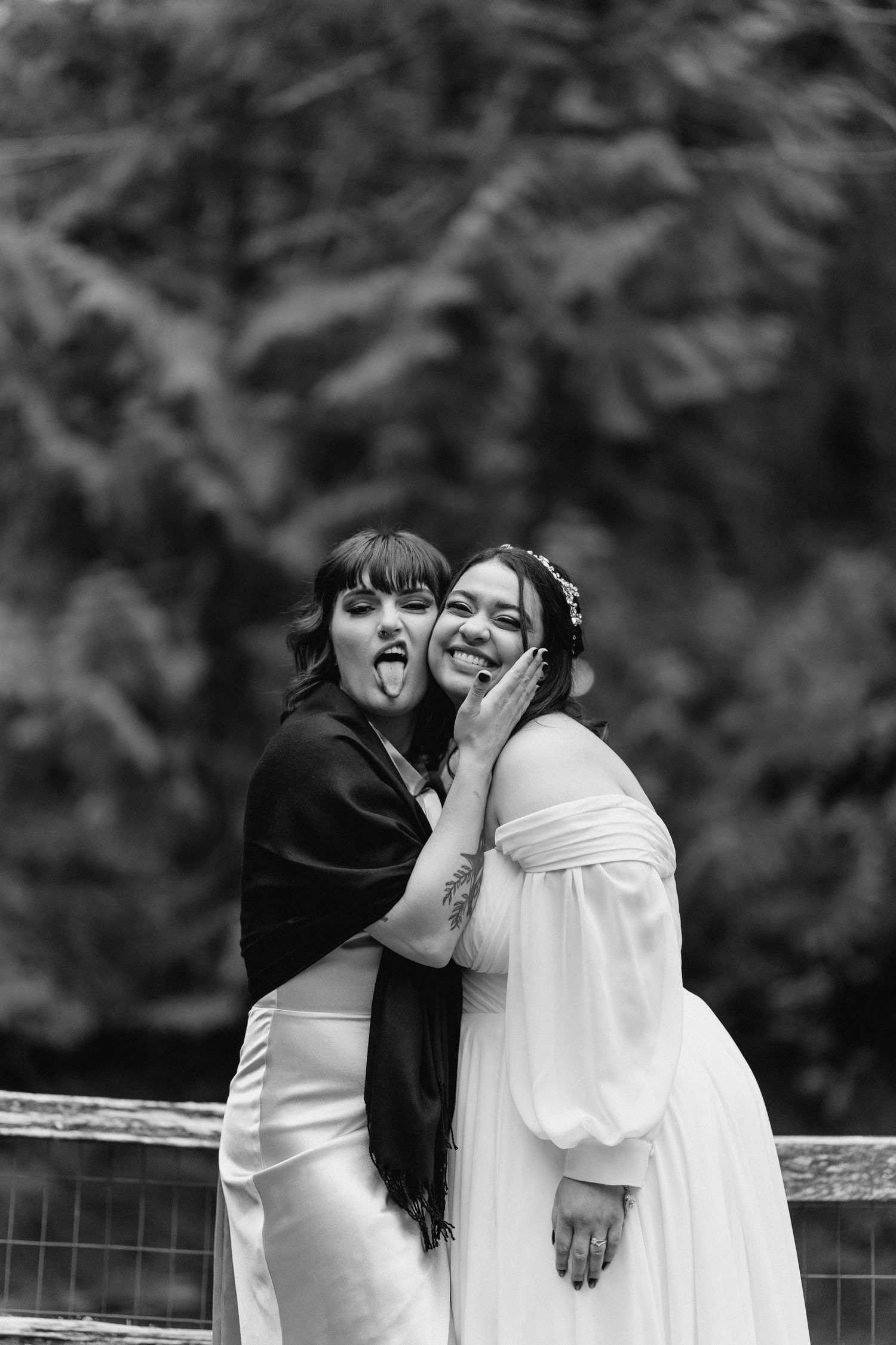 Playful bridesmaid sticks out tongue beside smiling bride in fun black and white wedding photo.