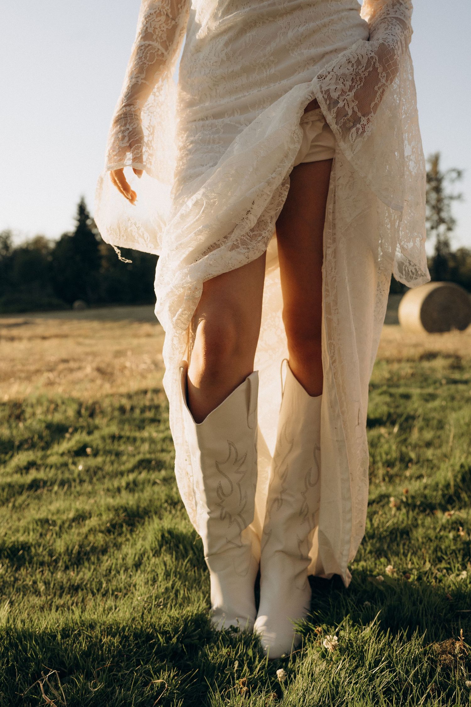Bride lifts lace wedding dress to reveal white cowboy boots in a sunlit field.