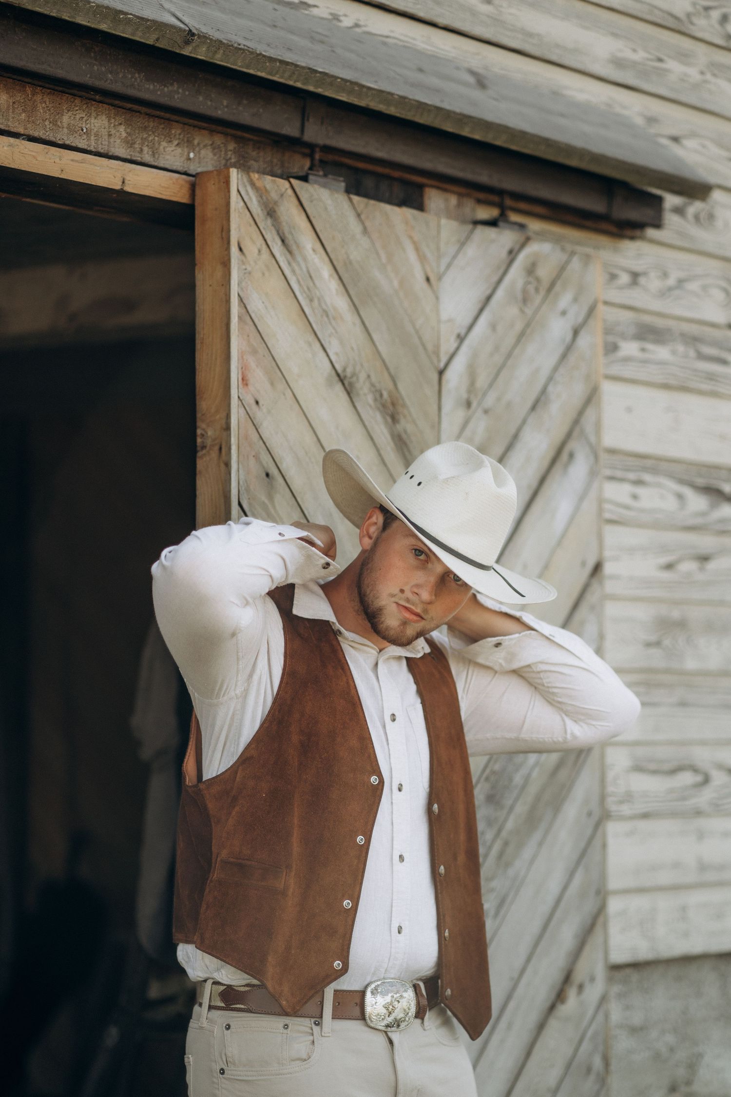 Groom in brown suede vest adjusting white cowboy hat outside rustic wooden barn.