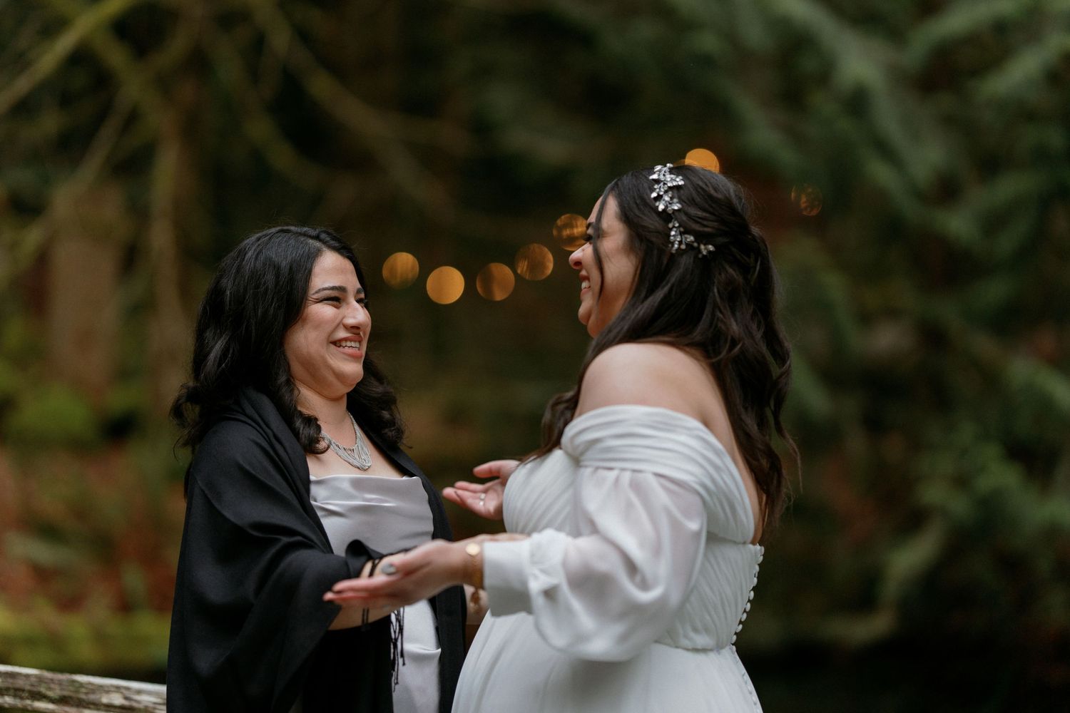 Two brides laughing and holding hands during outdoor wedding ceremony in forest.