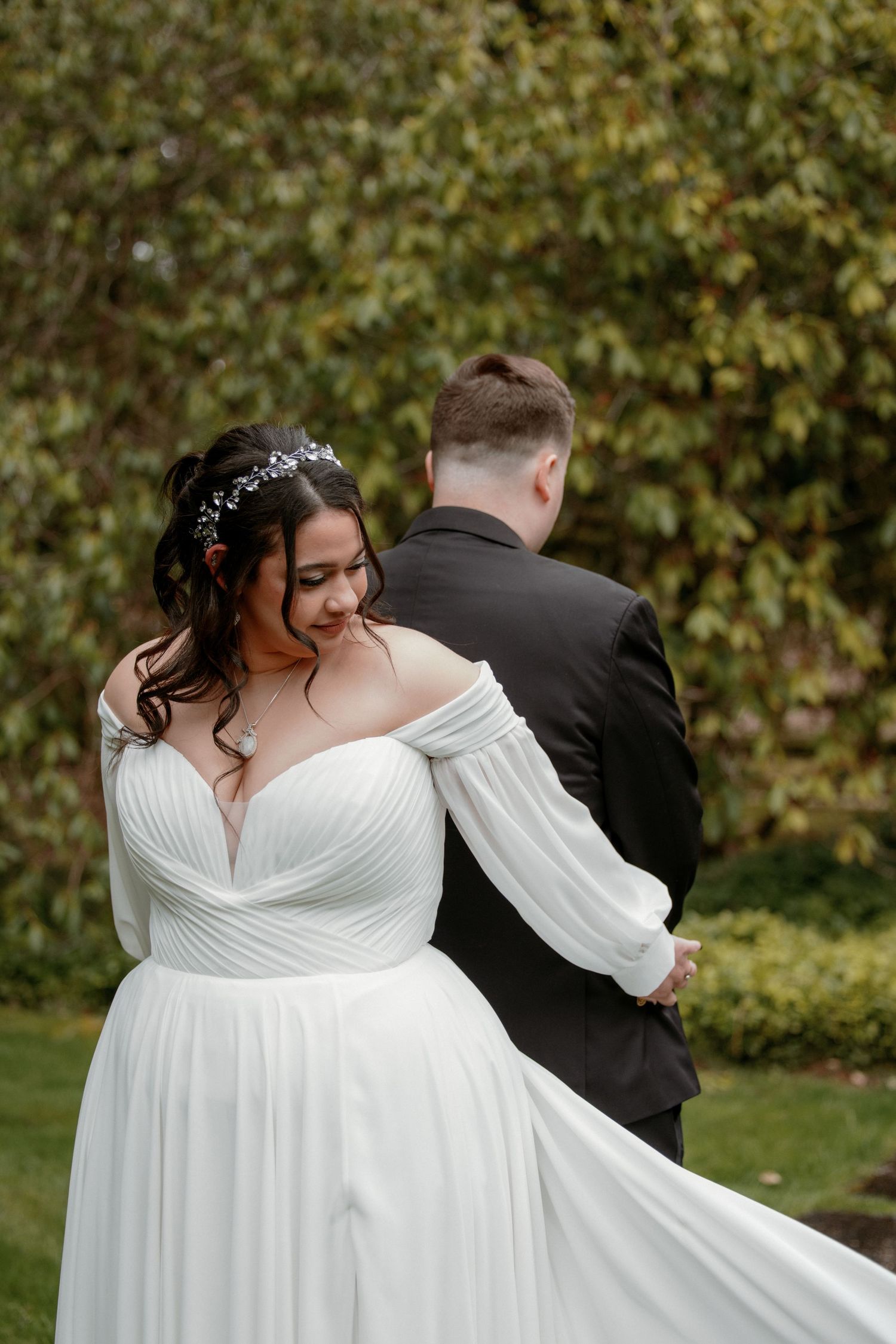 Bride in off-shoulder white gown embracing groom in black suit from behind in a garden setting.