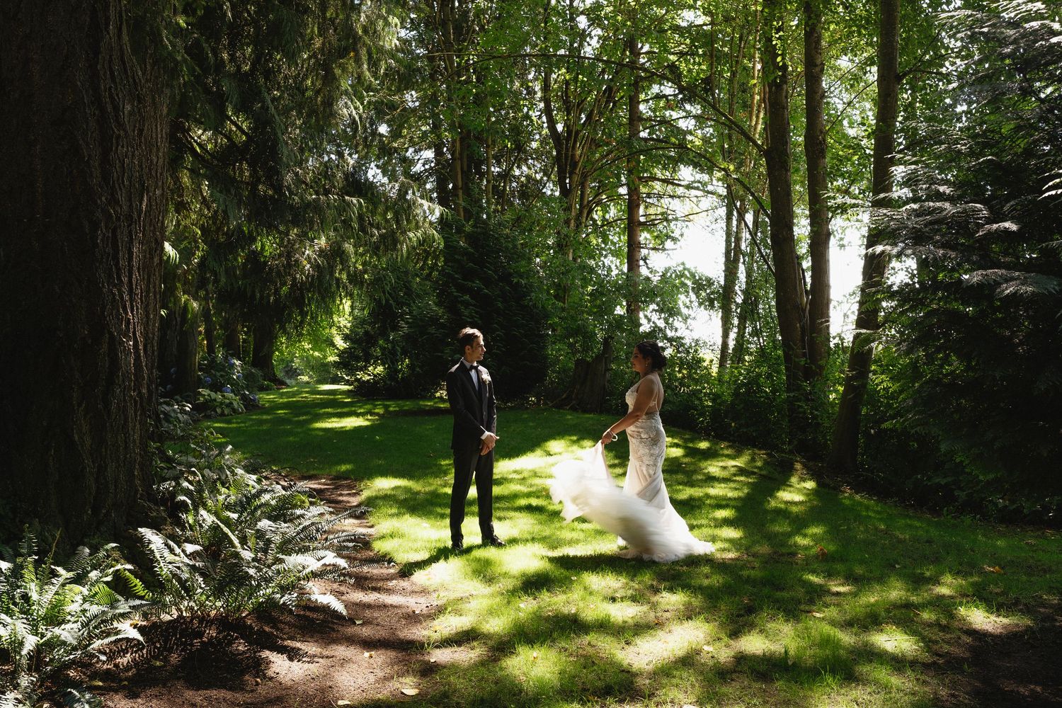 Bride in flowing white gown runs toward groom standing on a sunlit tree-lined garden path.