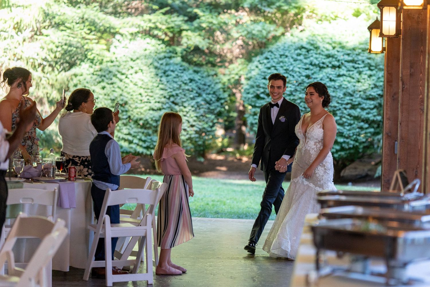Bride and groom make their grand entrance at an outdoor wedding reception venue surrounded by lush greenery.
