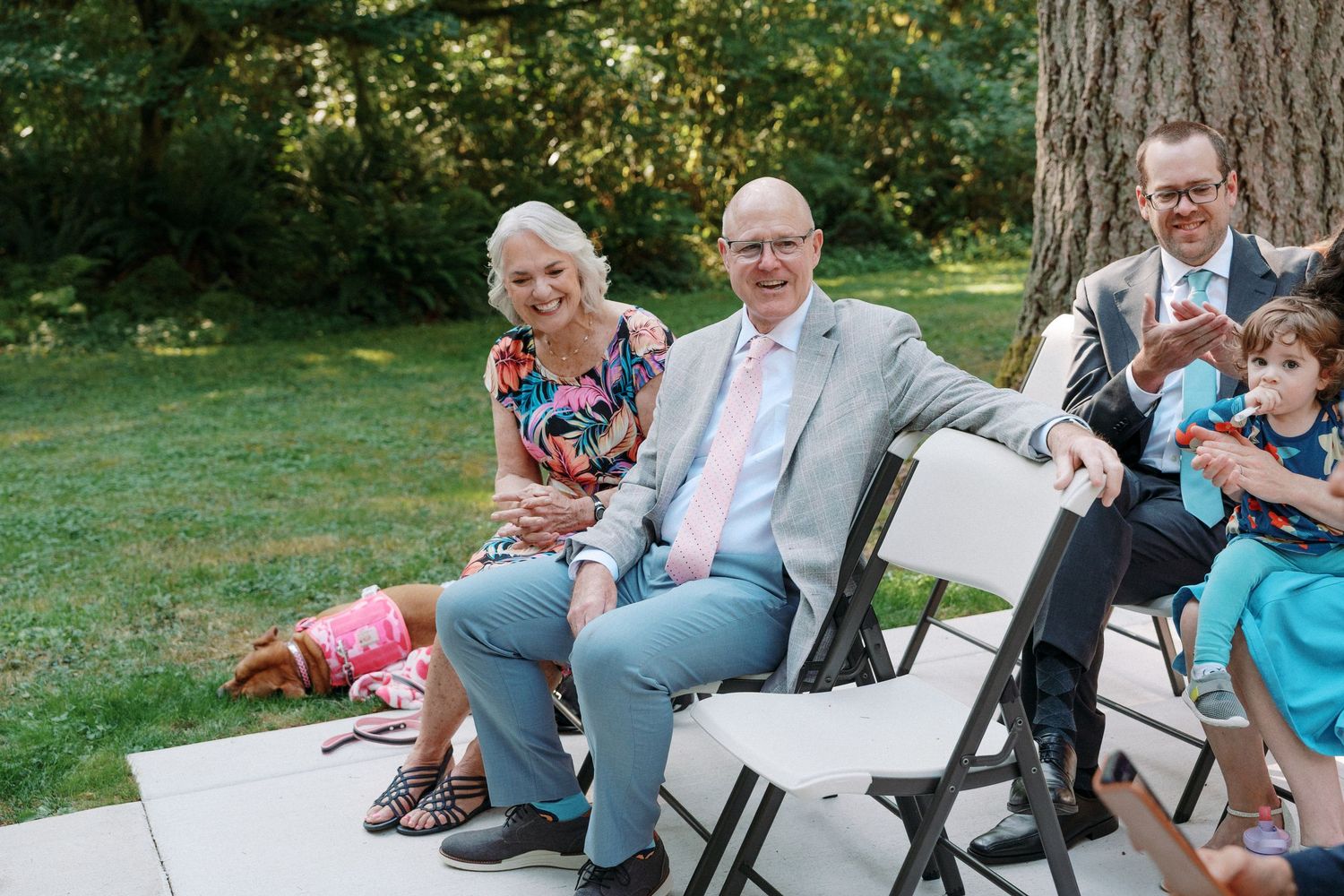 Elderly couple smiles warmly while seated at outdoor wedding ceremony on white folding chairs.