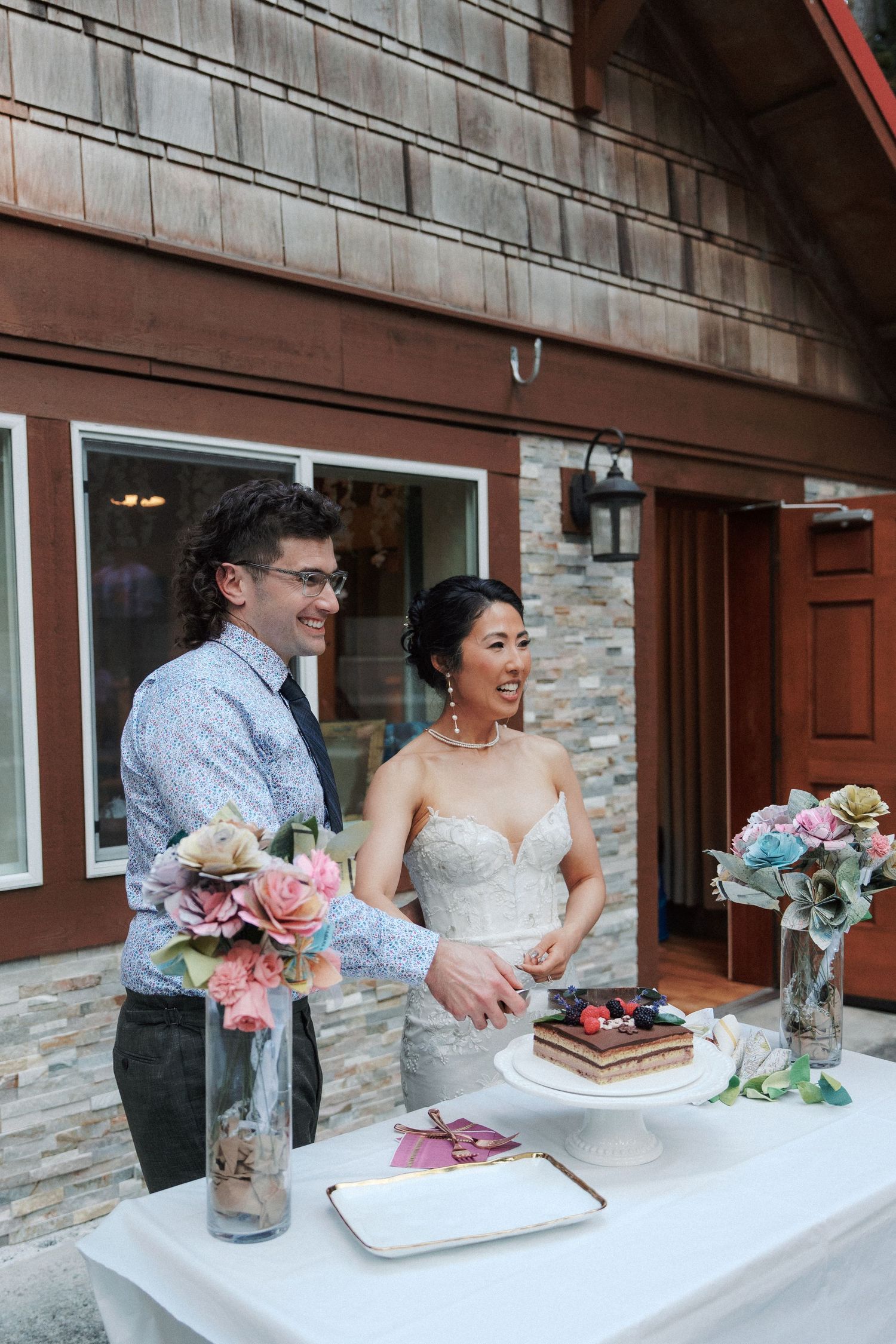 Bride and groom smiling while cutting wedding cake on outdoor table with floral arrangements.
