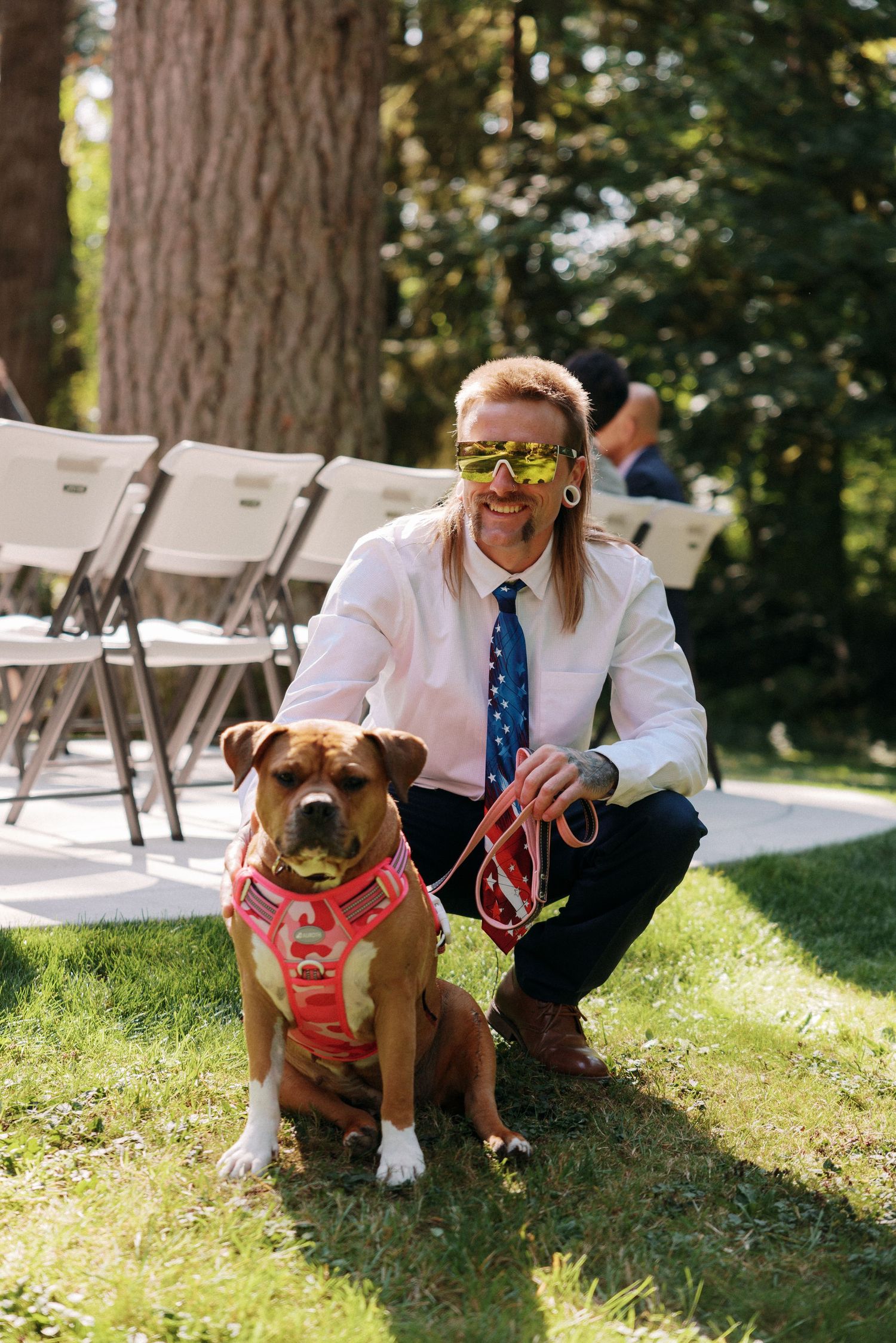 Man with long hair wearing star-shaped sunglasses and American flag tie posing with brown dog at outdoor wedding.