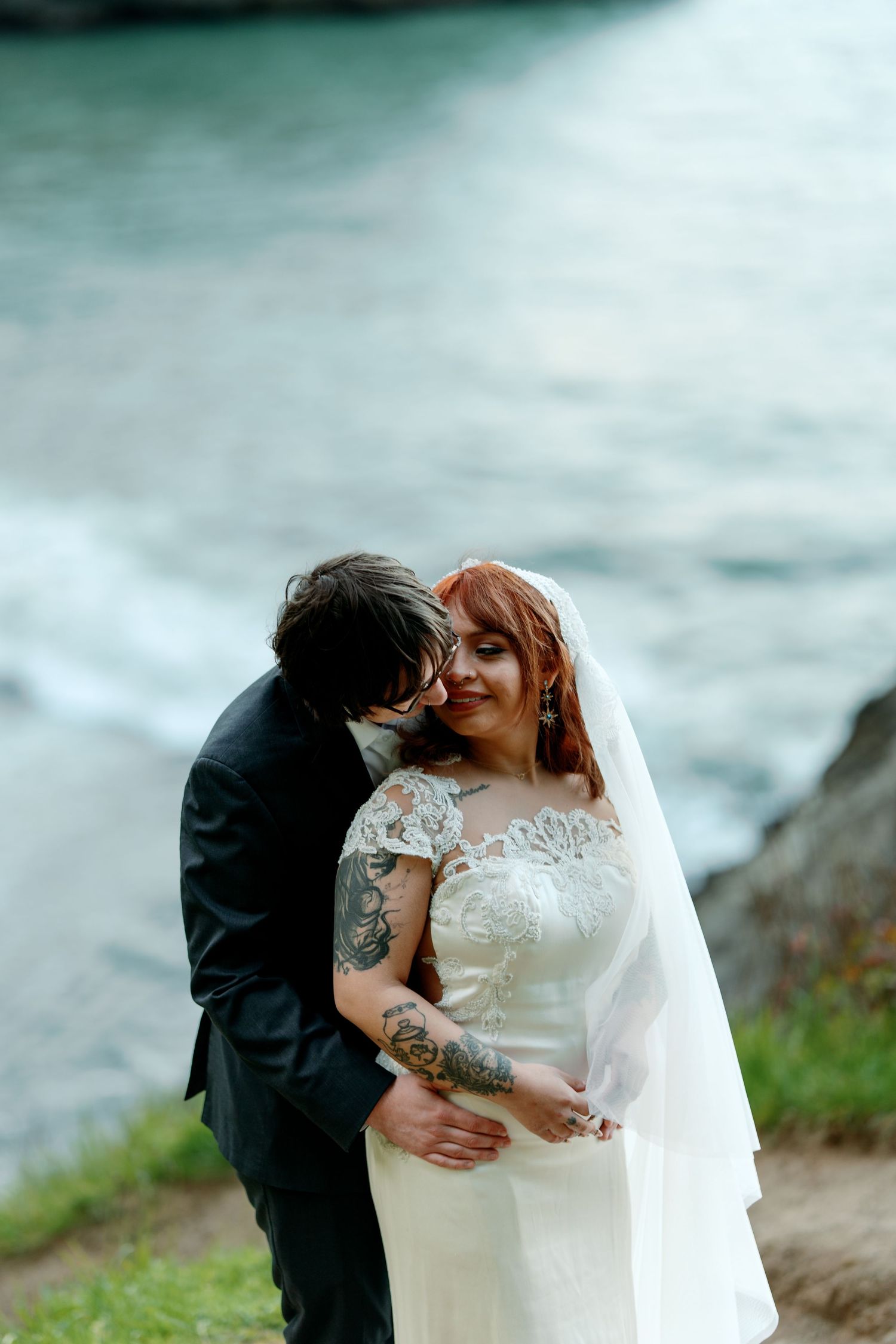 Tattooed redhead bride in lace wedding dress smiles as groom whispers in her ear by the ocean.