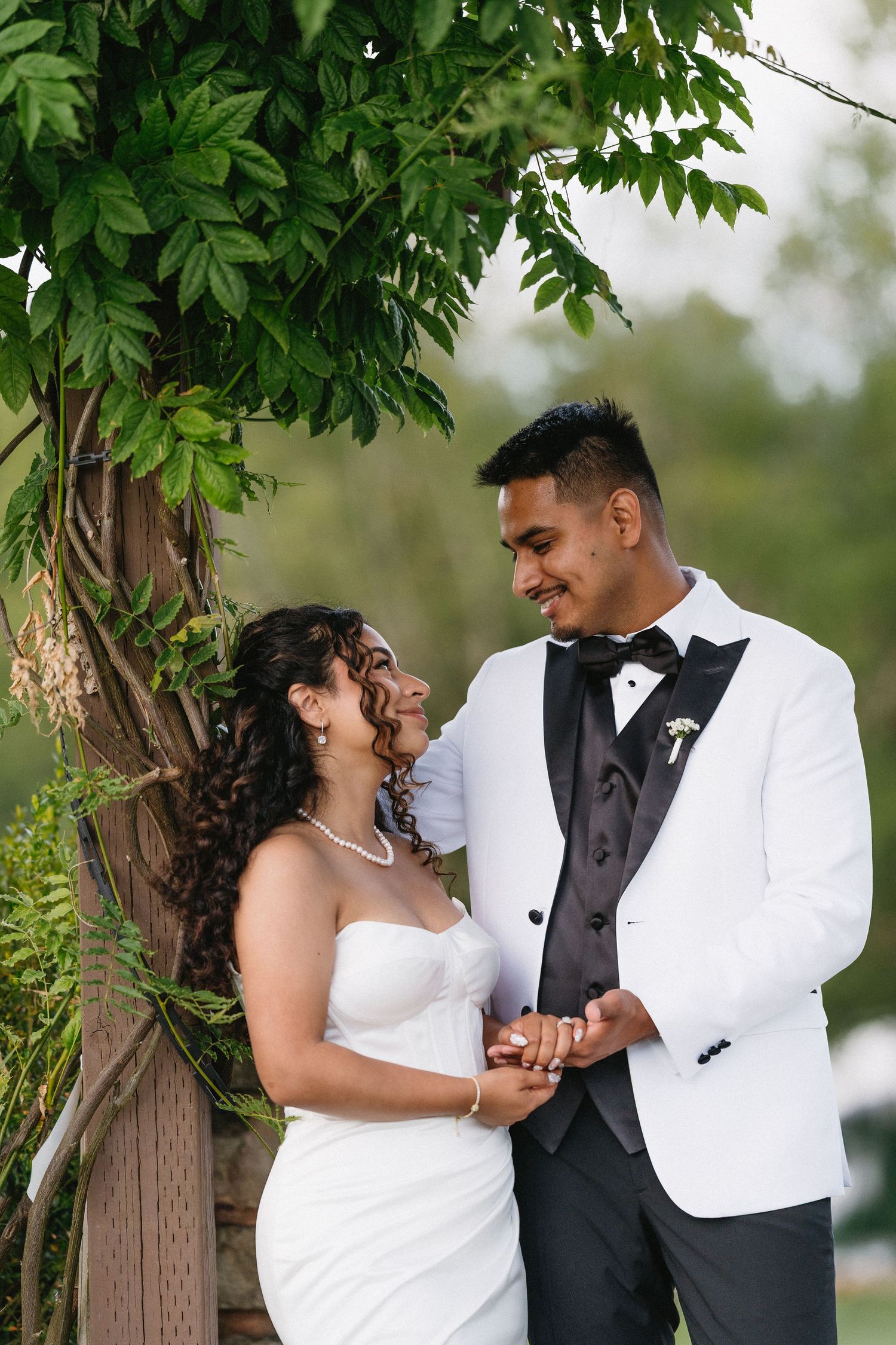Bride and groom share a joyful moment under a wooden arbor draped with green vines at outdoor wedding.