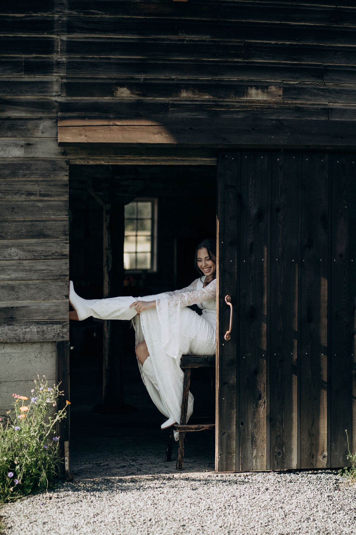 Joyful bride in white lace gown playfully kicking leg while seated in dark wooden barn doorway.