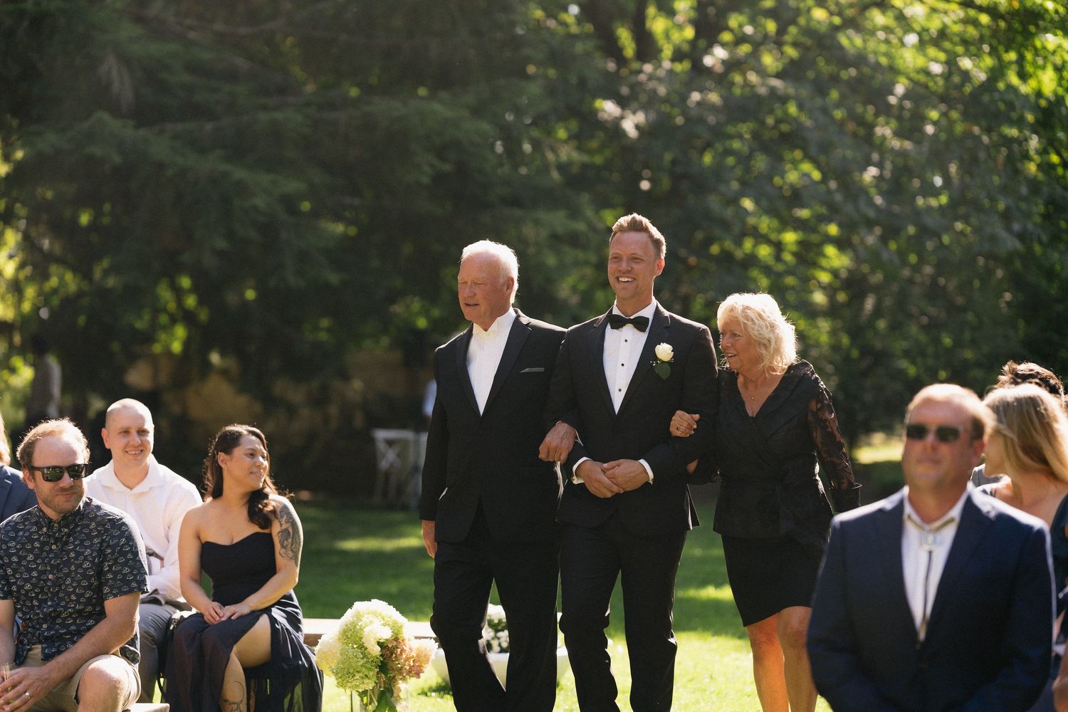 Groom in tuxedo walks down outdoor wedding aisle escorted by his parents in sunny garden setting.