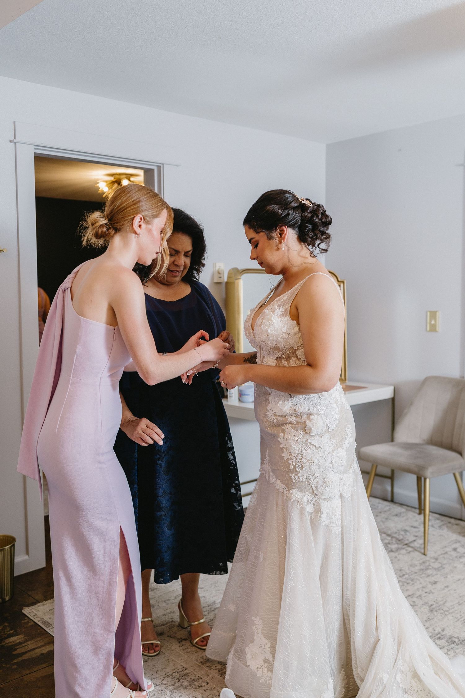 Bridesmaid and guest helping bride in lace wedding dress with final preparations indoors.