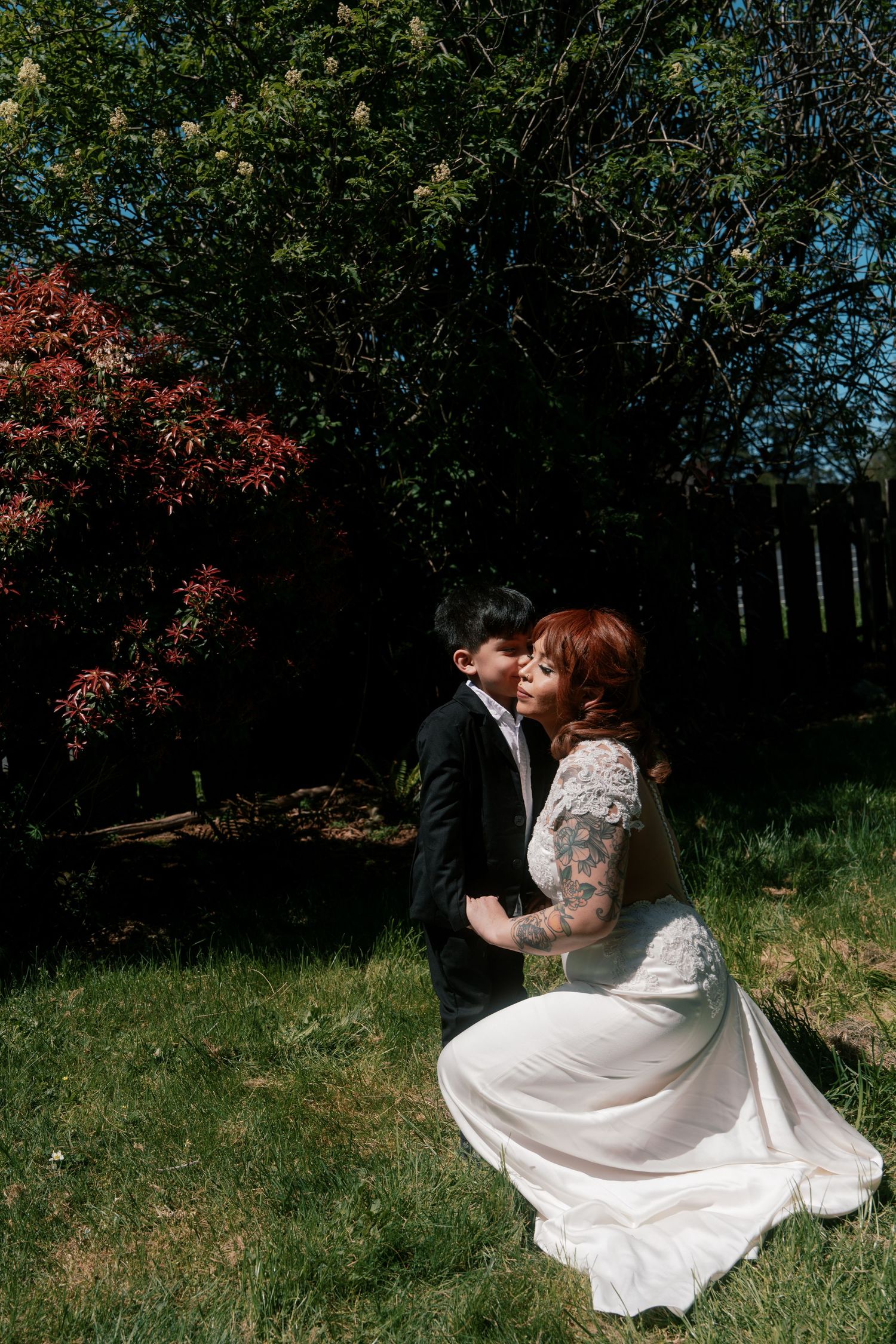 Bride in lace gown with floral tattoos shares a romantic kiss with groom in sunny backyard garden.
