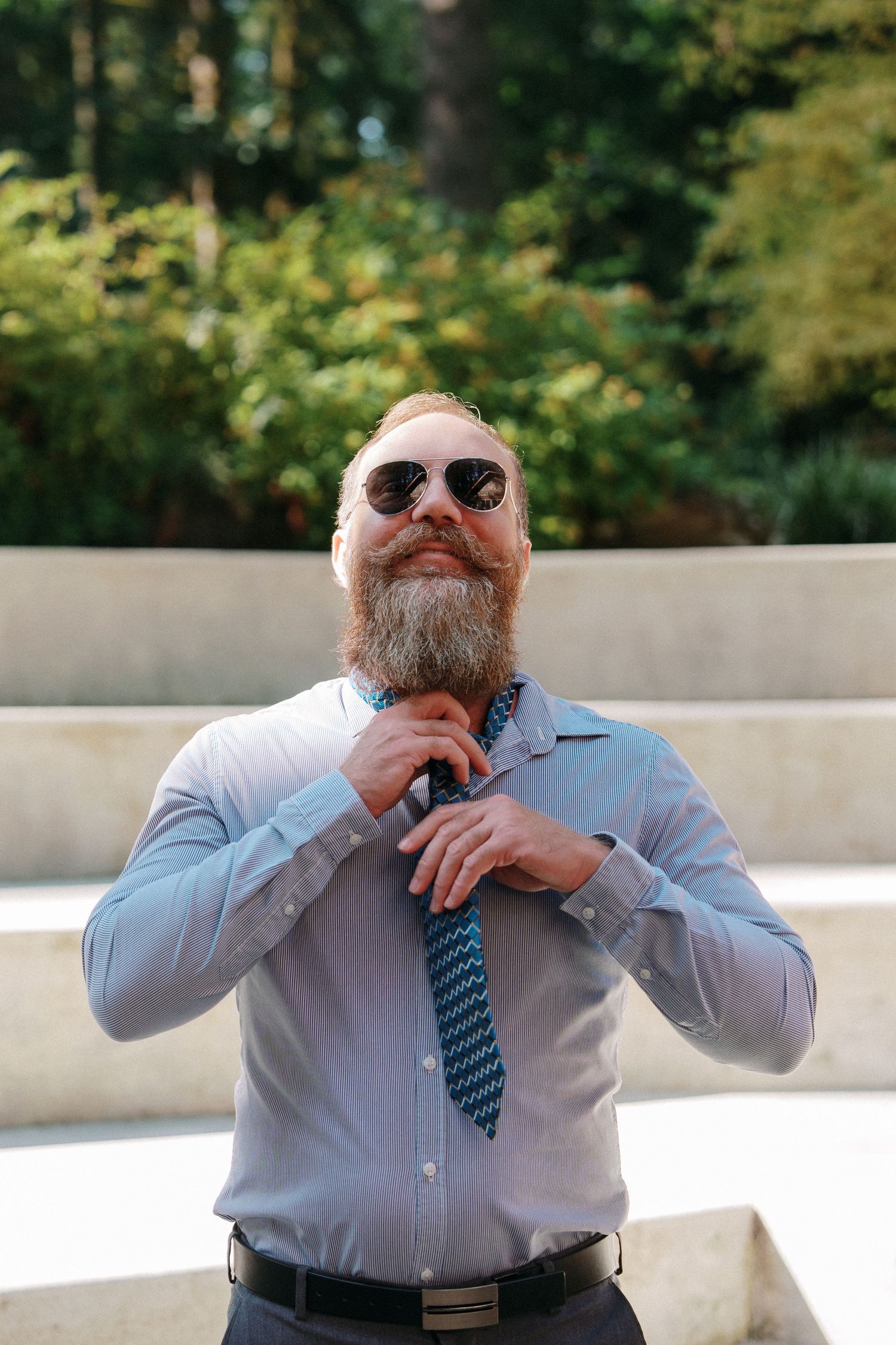 A bearded man in sunglasses adjusts his blue tie while standing outdoors in a striped dress shirt.