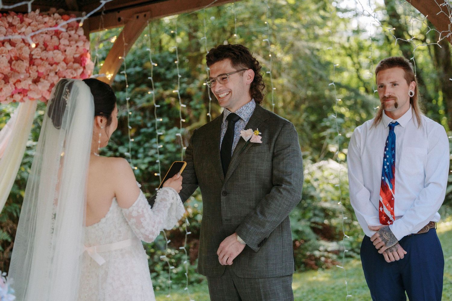 Groom smiles warmly at bride during intimate outdoor wedding ceremony beneath floral-decorated wooden pergola.